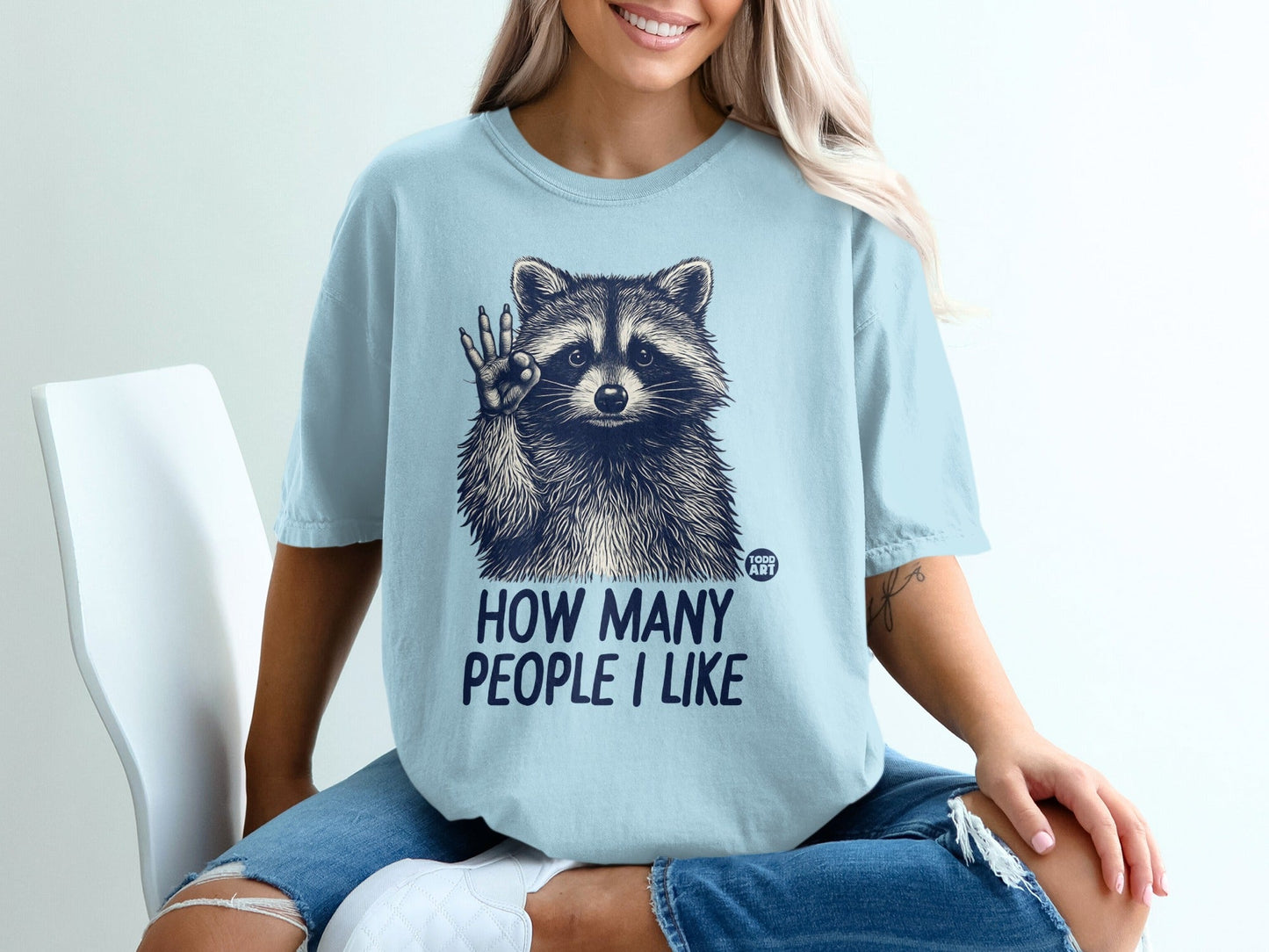 Light blue t-shirt with raccoon illustration and text "HOW MANY PEOPLE I LIKE".