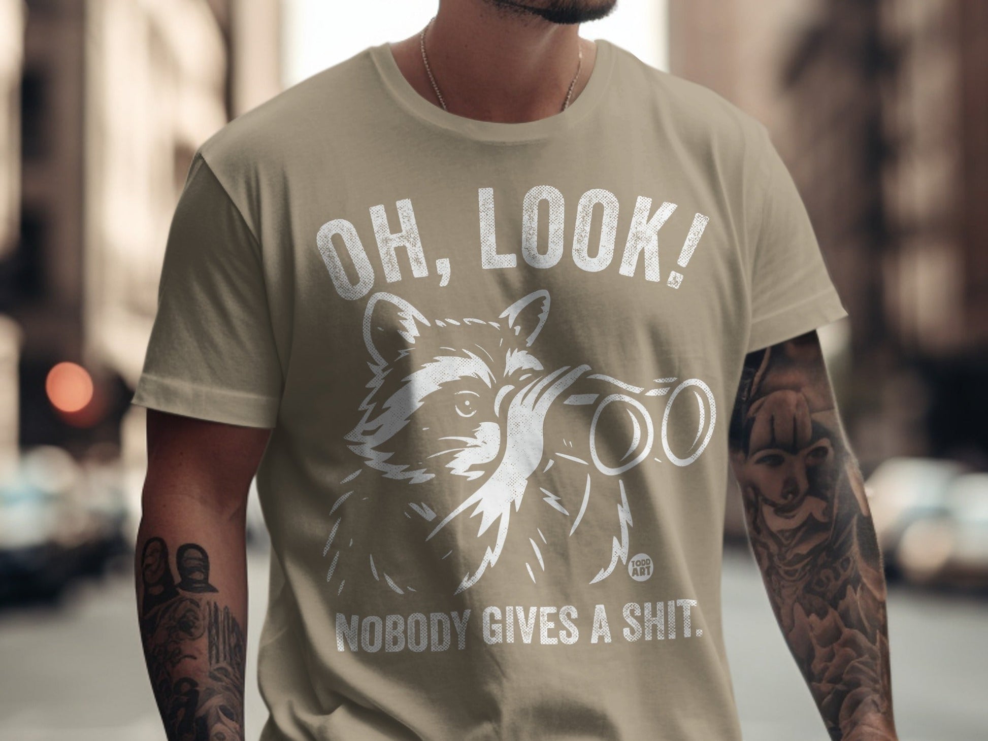 Tan t-shirt with raccoon looking through binoculars and text "OH, LOOK! NOBODY GIVES A SHIT."