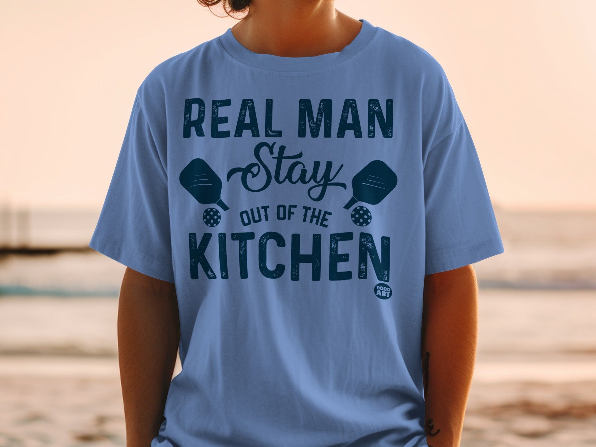 Blue t-shirt with "Real Man Stay Out of the Kitchen" text and pickleballs.