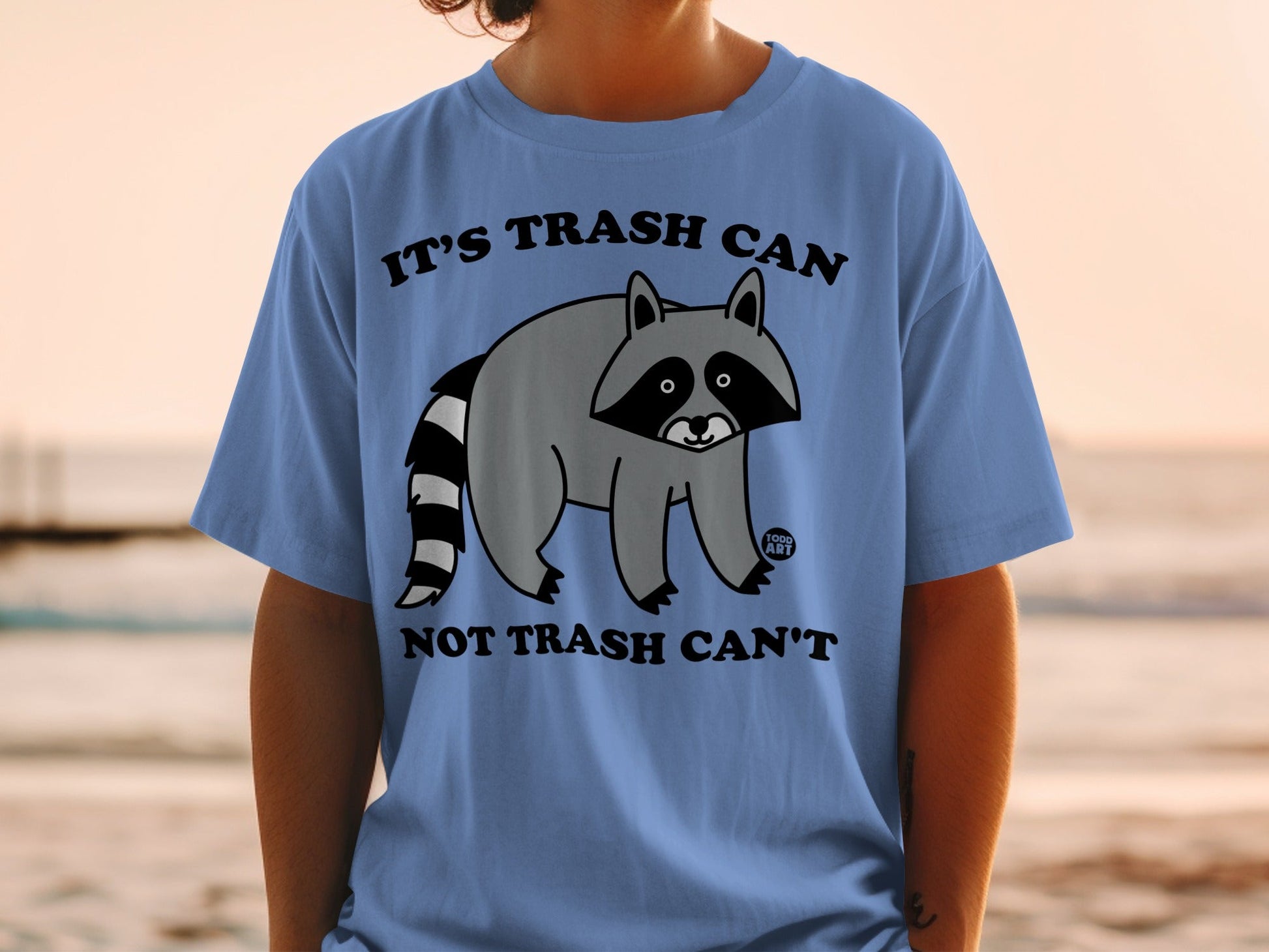 Blue t-shirt with raccoon illustration and text "IT'S TRASH CAN NOT TRASH CAN'T".