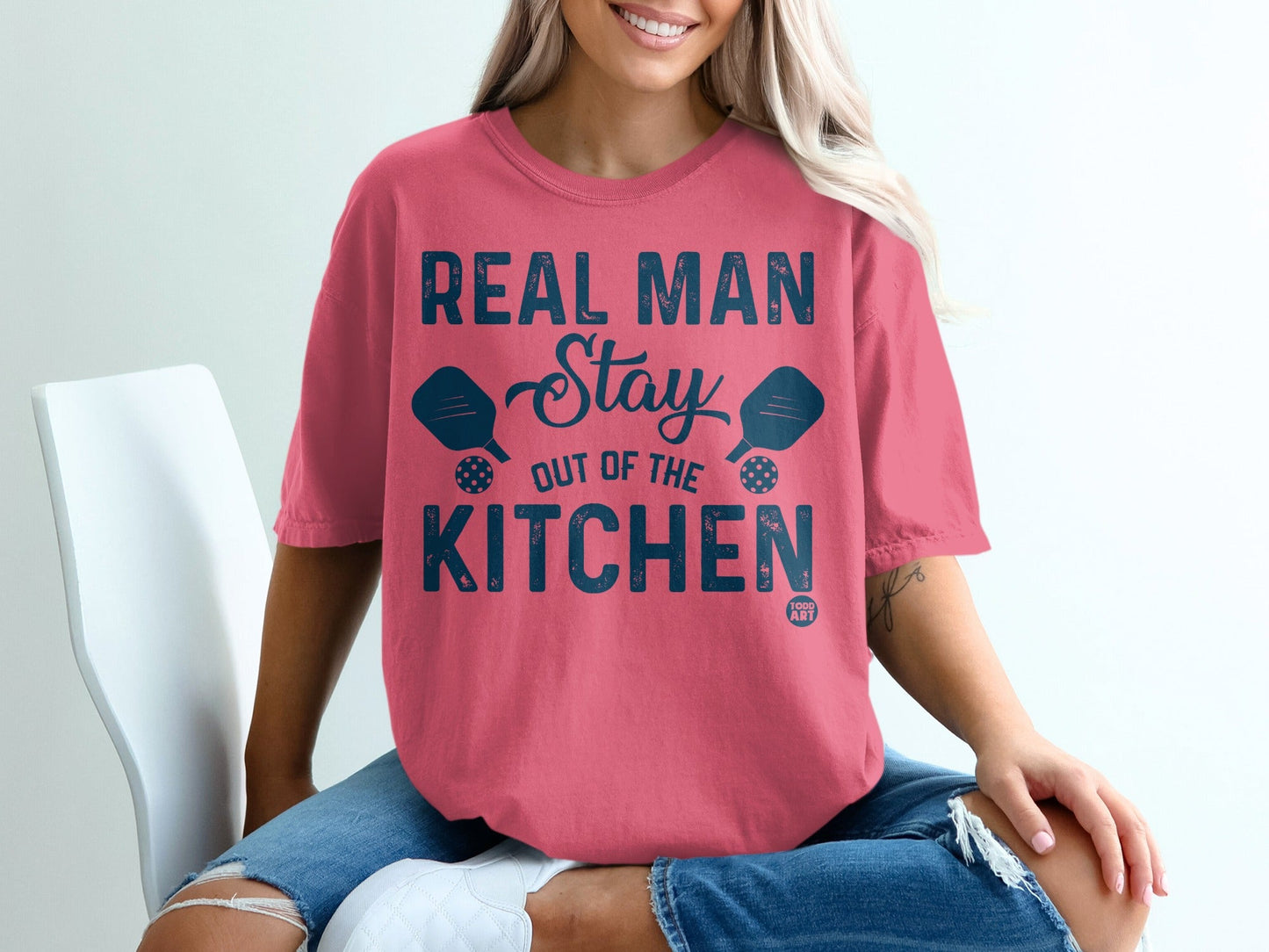 Coral t-shirt with "Real Man Stay Out of the Kitchen" text and pickleball paddles.