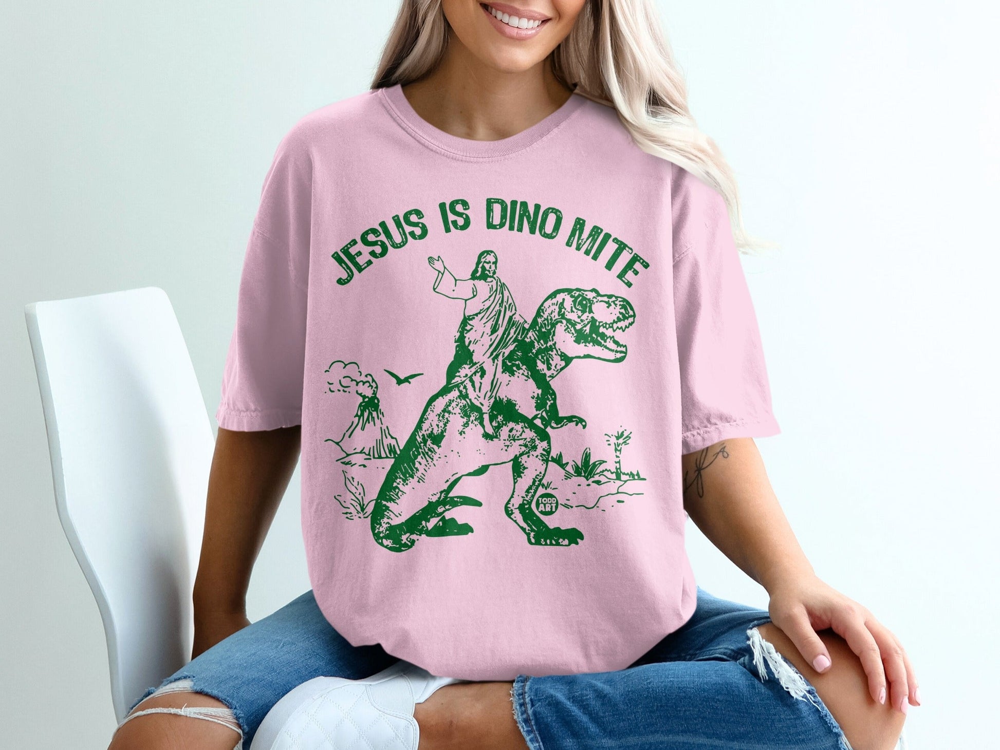 Pink t-shirt with green graphic of Jesus riding a dinosaur.