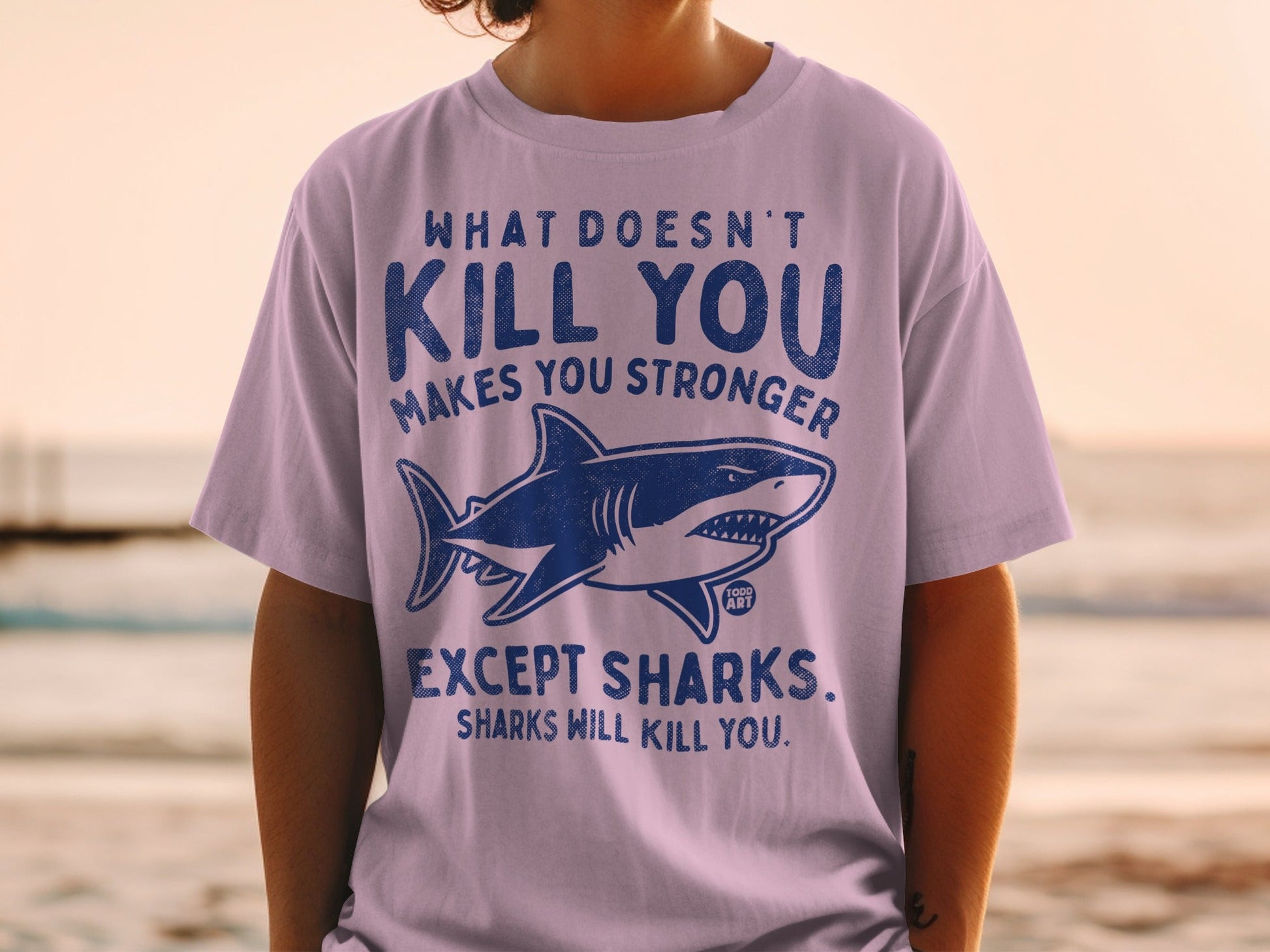 Light purple t-shirt with shark graphic and humorous text "What Doesn't Kill You Makes You Stronger Except Sharks."