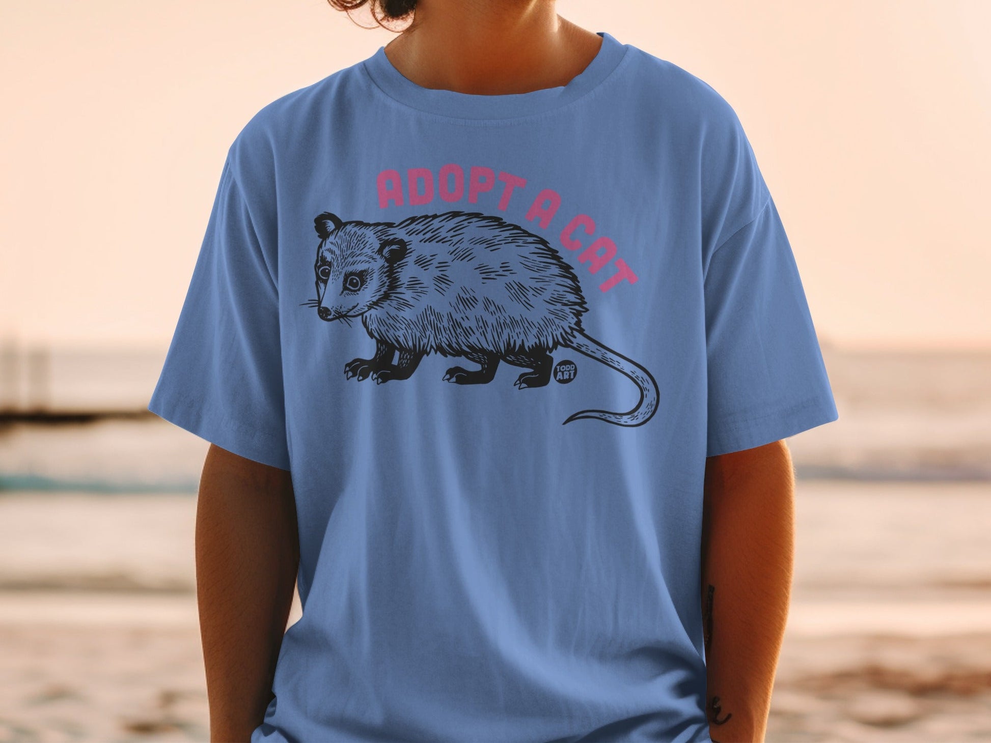 Blue t-shirt with pink text "ADOPT A CAT" and a drawing of an opossum.