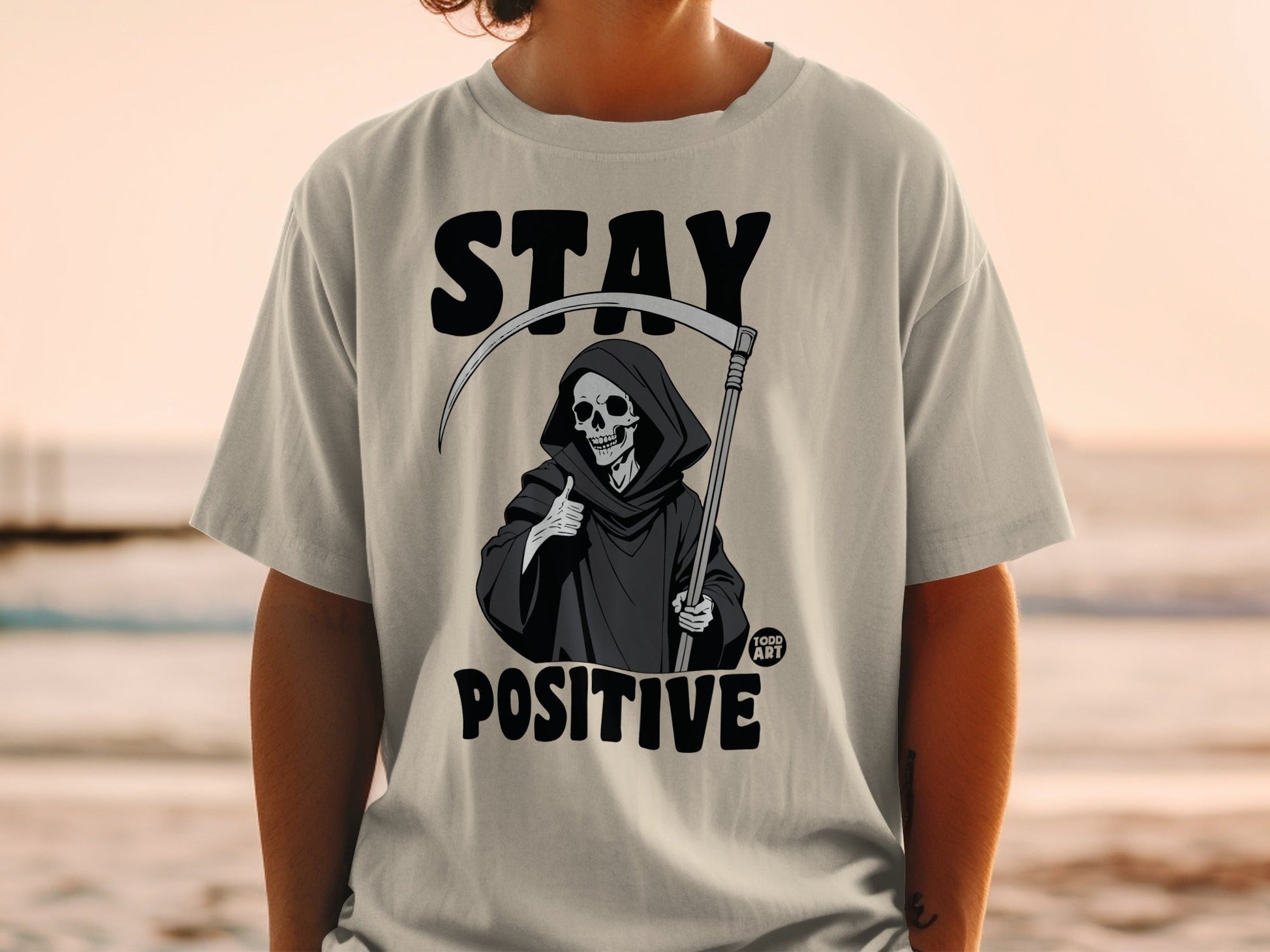 Grim reaper giving thumbs up on t-shirt with "STAY POSITIVE" text.