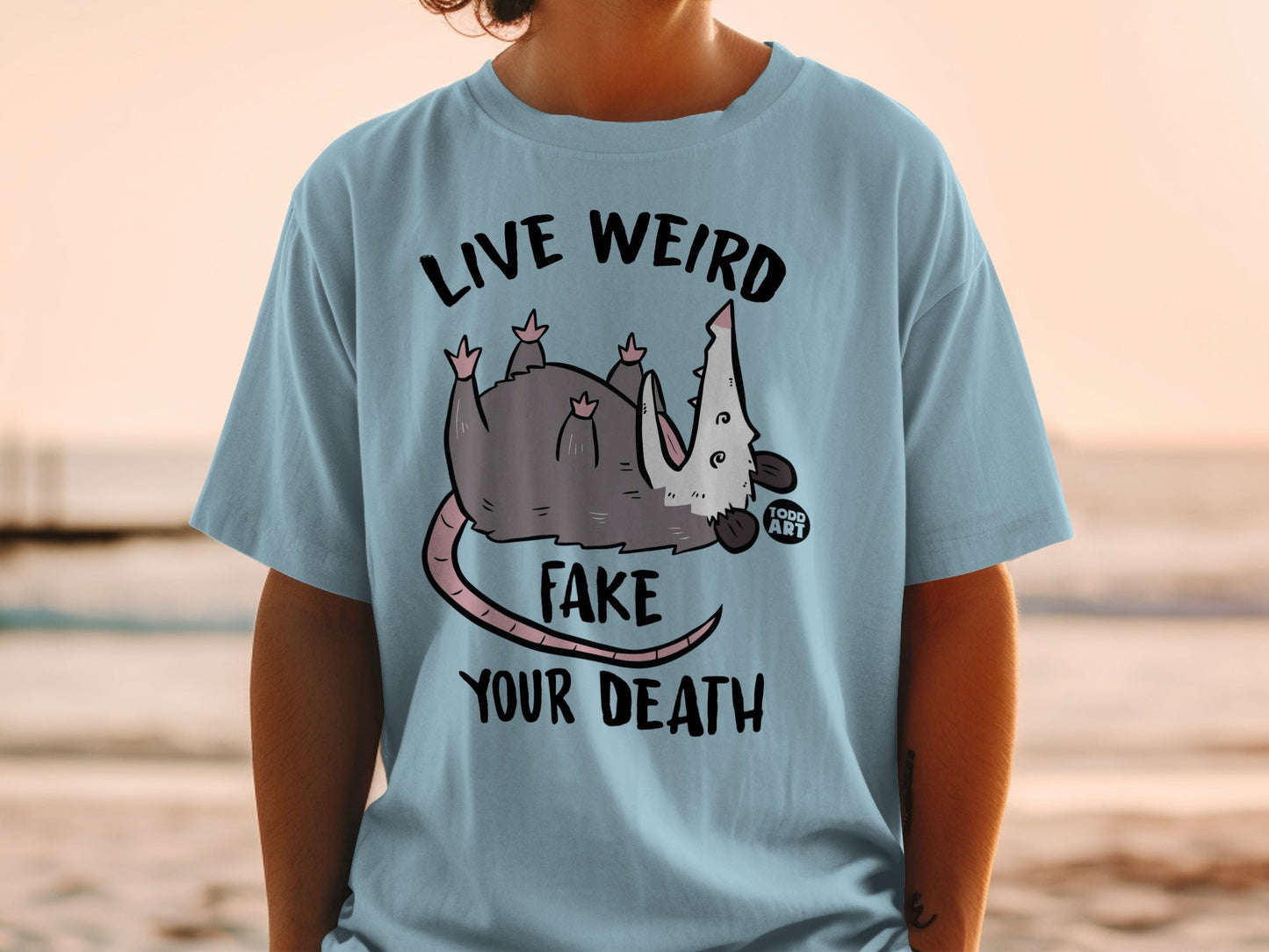 Blue t-shirt with opossum playing dead and text "LIVE WEIRD FAKE YOUR DEATH".