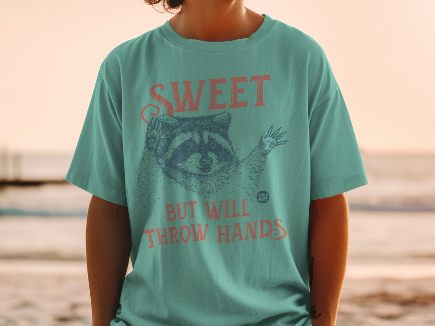Teal t-shirt with raccoon graphic and "SWEET BUT WILL THROW HANDS" text.