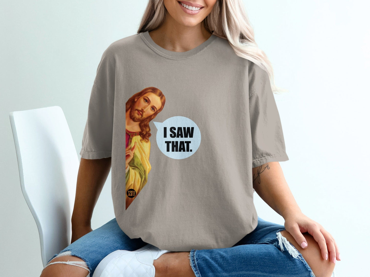 Woman wearing a tan t-shirt with Jesus looking from the side saying "I SAW THAT."
