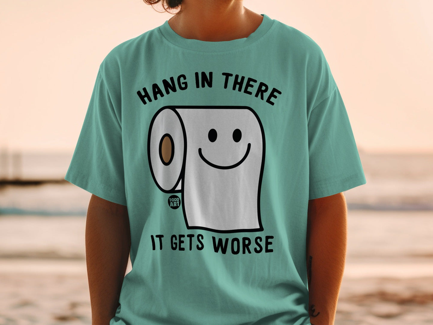 Teal t-shirt with a smiling toilet paper roll and text "HANG IN THERE IT GETS WORSE".