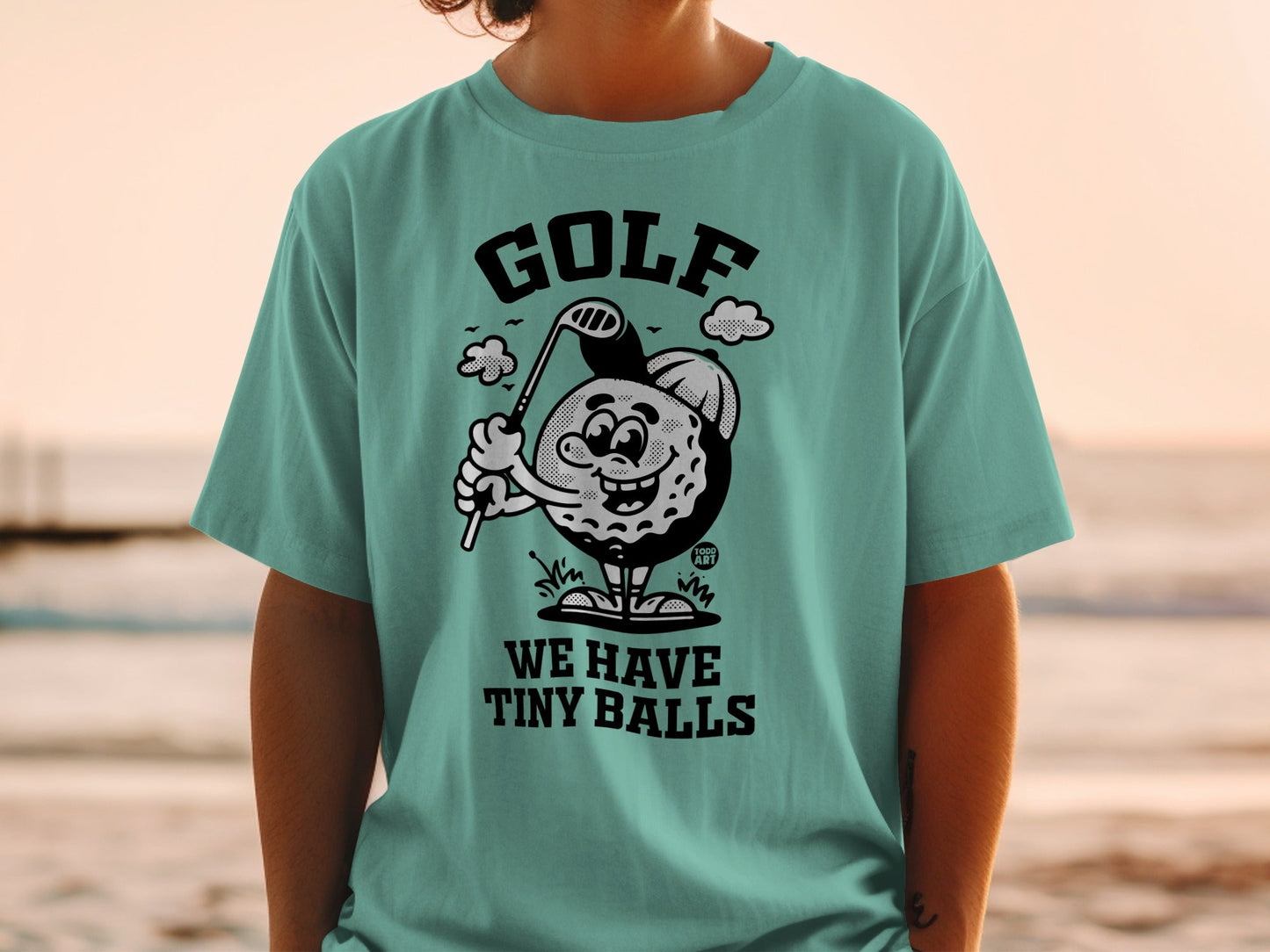 T-shirt featuring a cartoon golf ball character swinging a club with the text "WE HAVE TINY BALLS."
