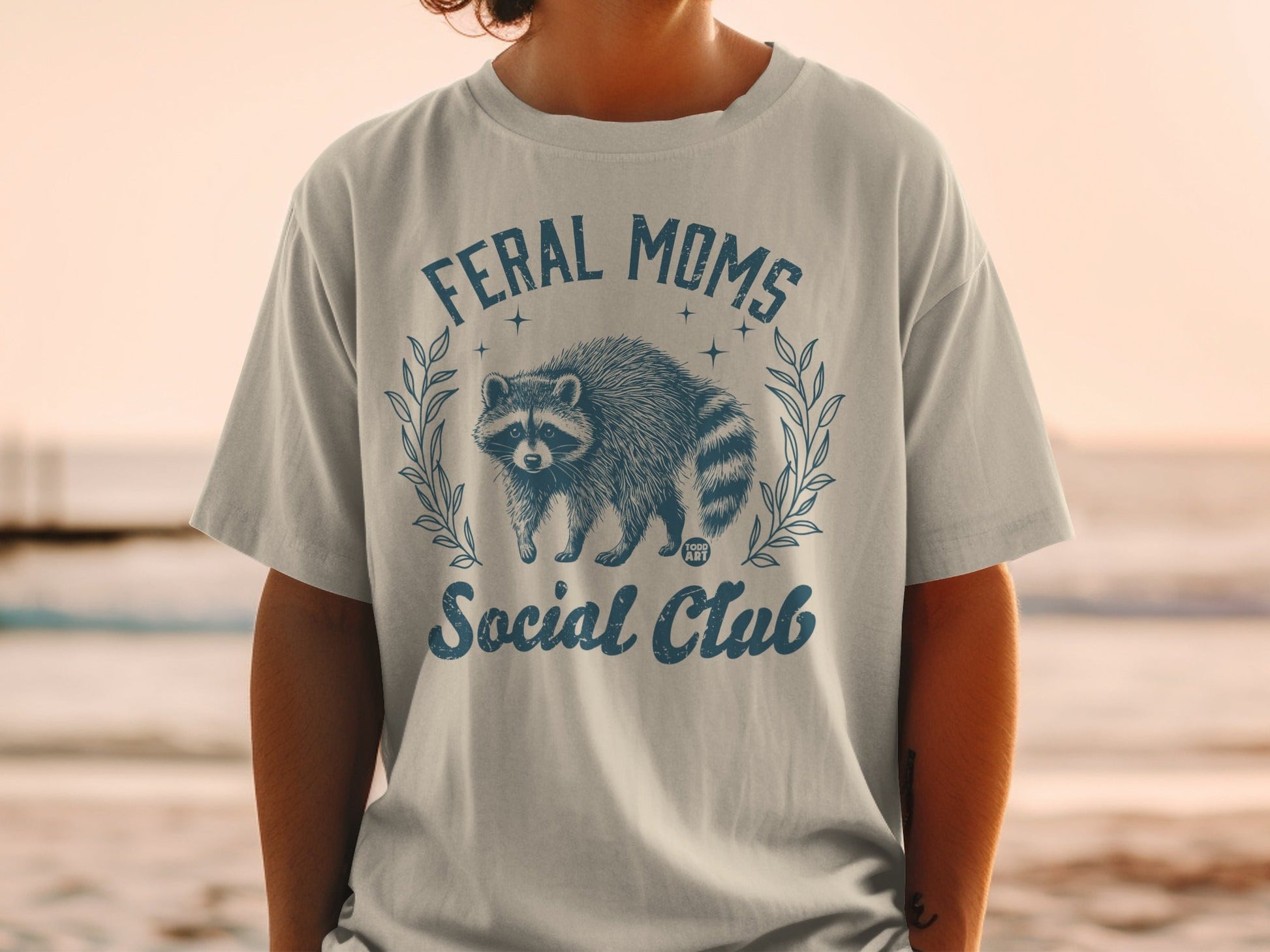 Feral Moms Social Club text with raccoon graphic on a t-shirt.