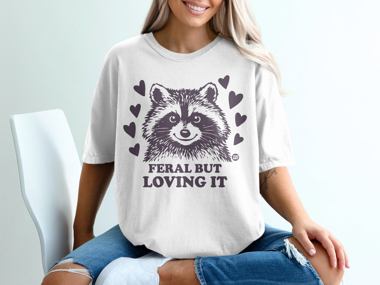 White t-shirt with smiling raccoon graphic and text "Feral but loving it".