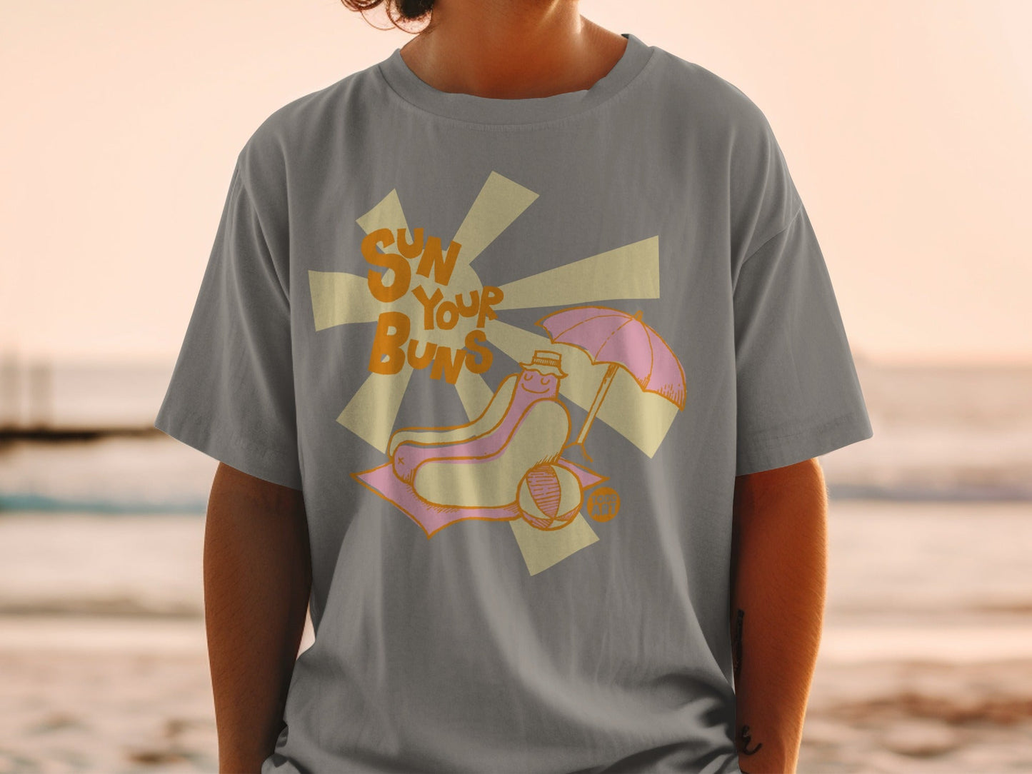 T-shirt with a hot dog wearing a hat under an umbrella on a beach.
