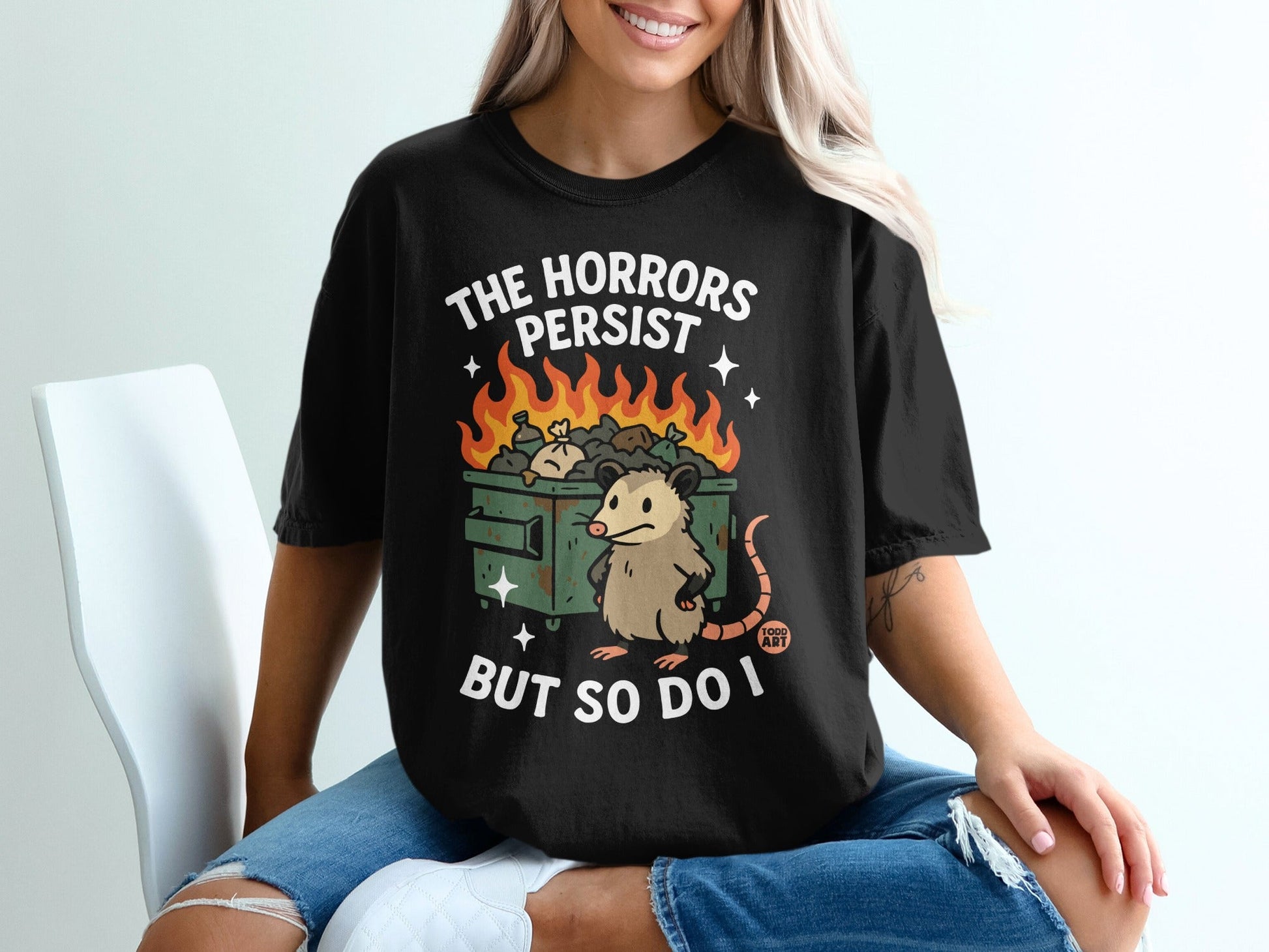T-shirt with possum sitting by burning trash can, text reads "The Horrors Persist But So Do I".