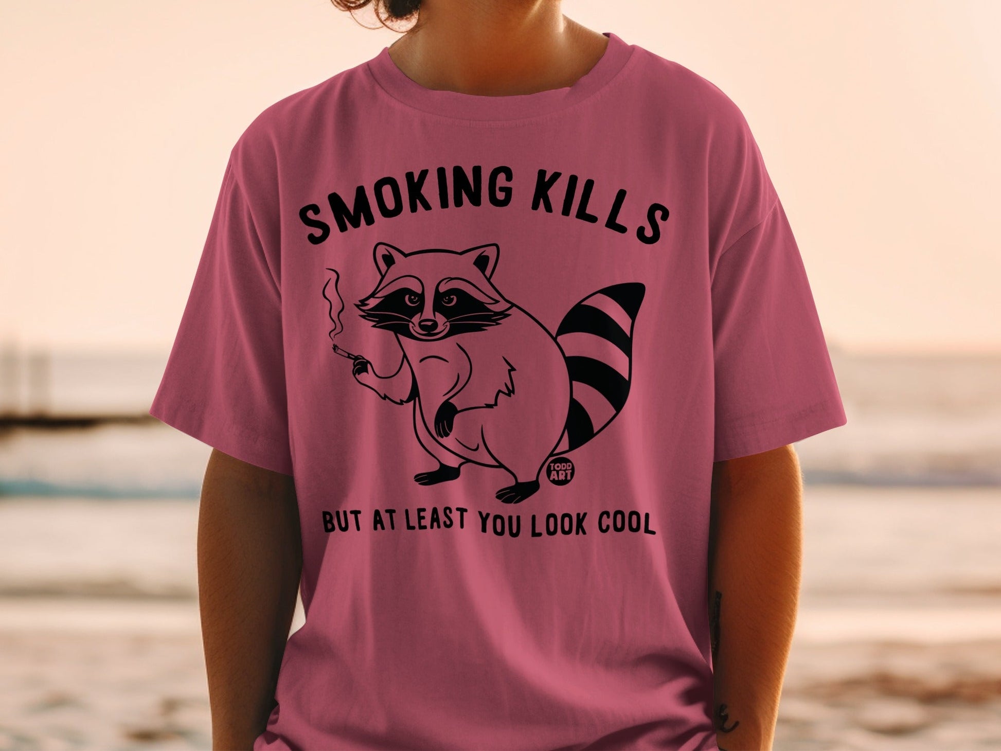 Pink t-shirt with raccoon smoking, reads "SMOKING KILLS BUT AT LEAST YOU LOOK COOL".