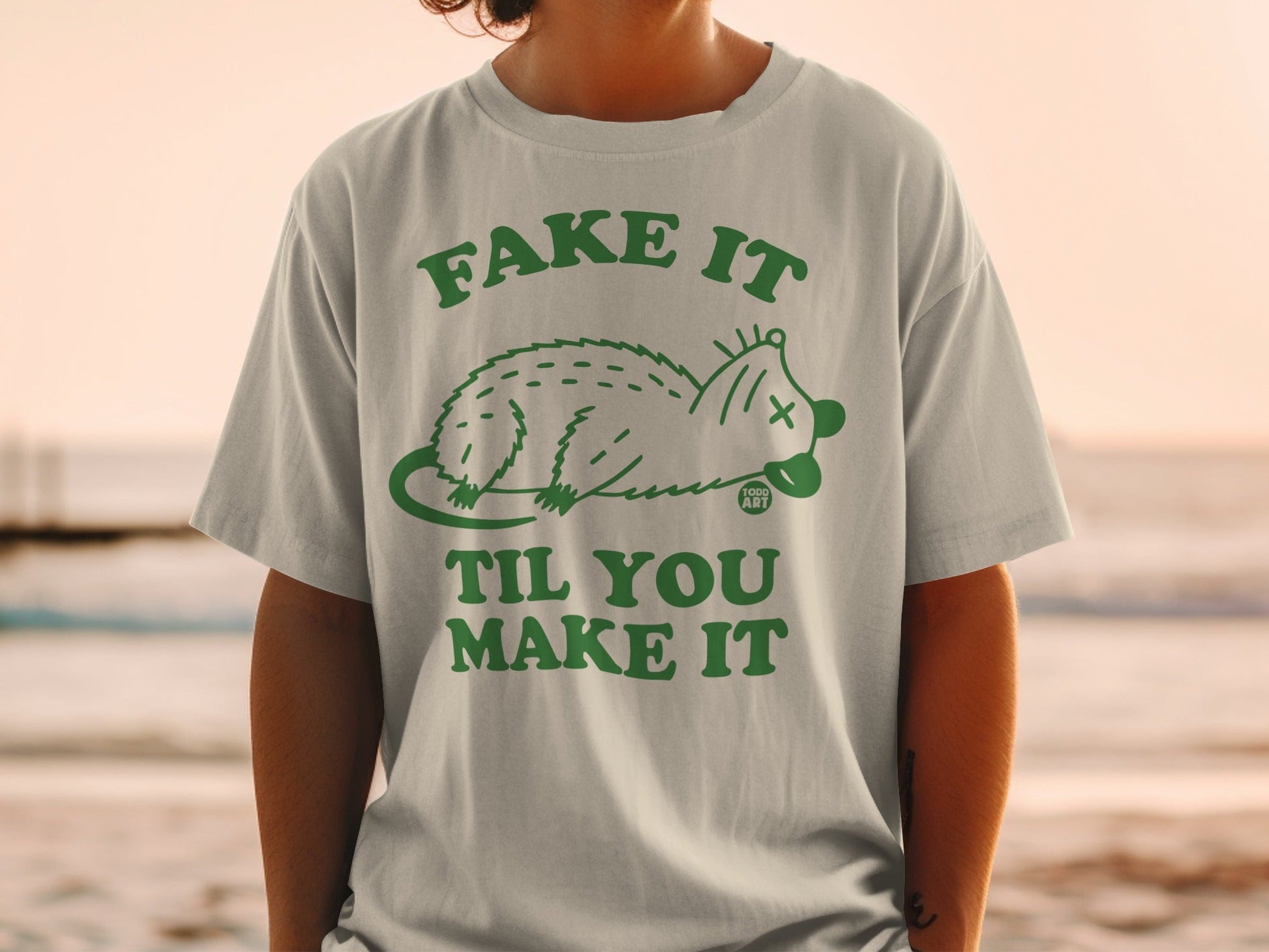 Light grey t-shirt with green text "FAKE IT TIL YOU MAKE IT" and a cartoon possum.