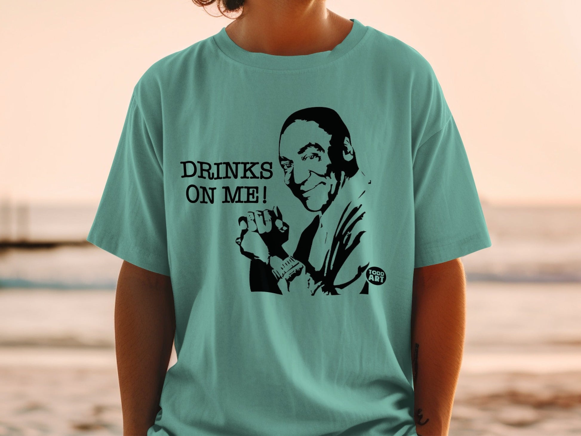 Teal t-shirt with a black graphic of a man saying "Drinks on me!".
