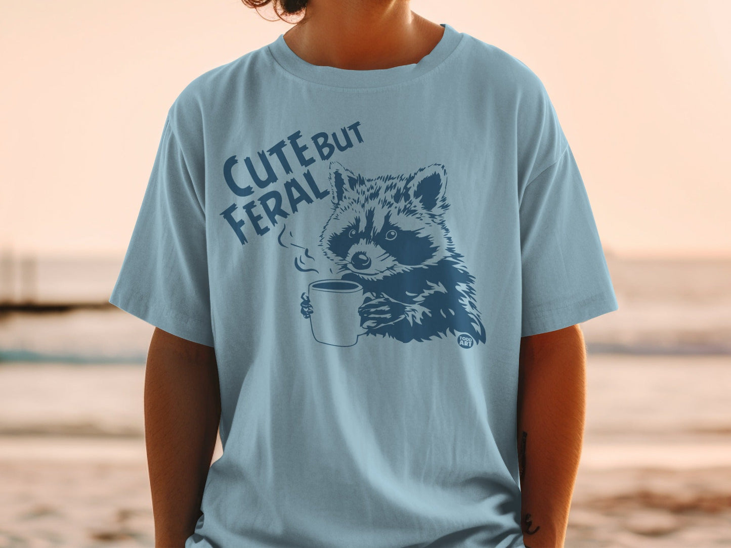 T-shirt with dark blue graphic of raccoon holding coffee cup, text "CUTE BUT FERAL".