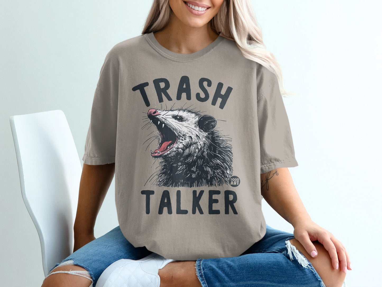 Trash Talker opossum graphic on a tan t-shirt worn by a woman.