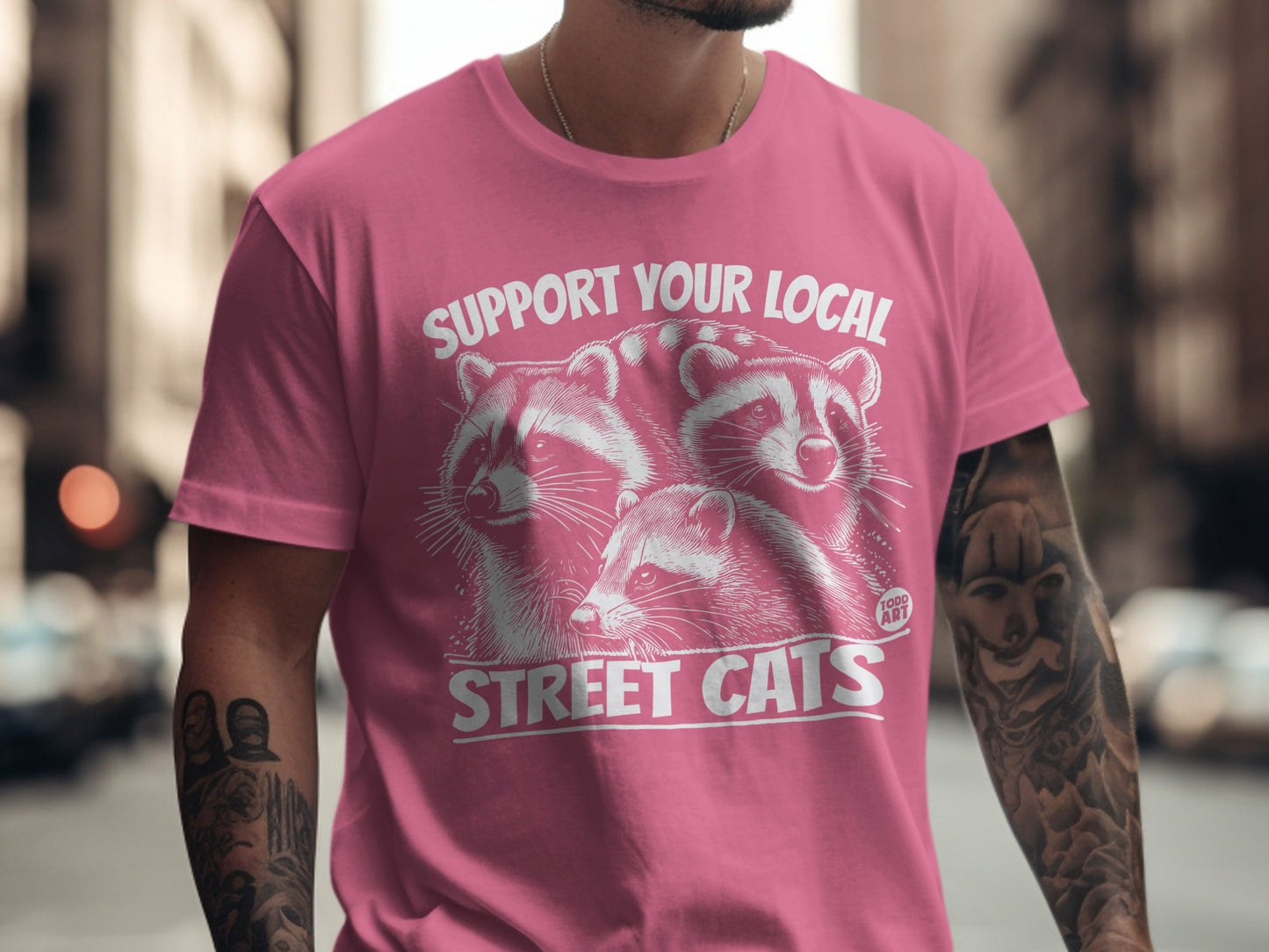 Pink t-shirt with graphic of three raccoons and text "SUPPORT YOUR LOCAL STREET CATS".