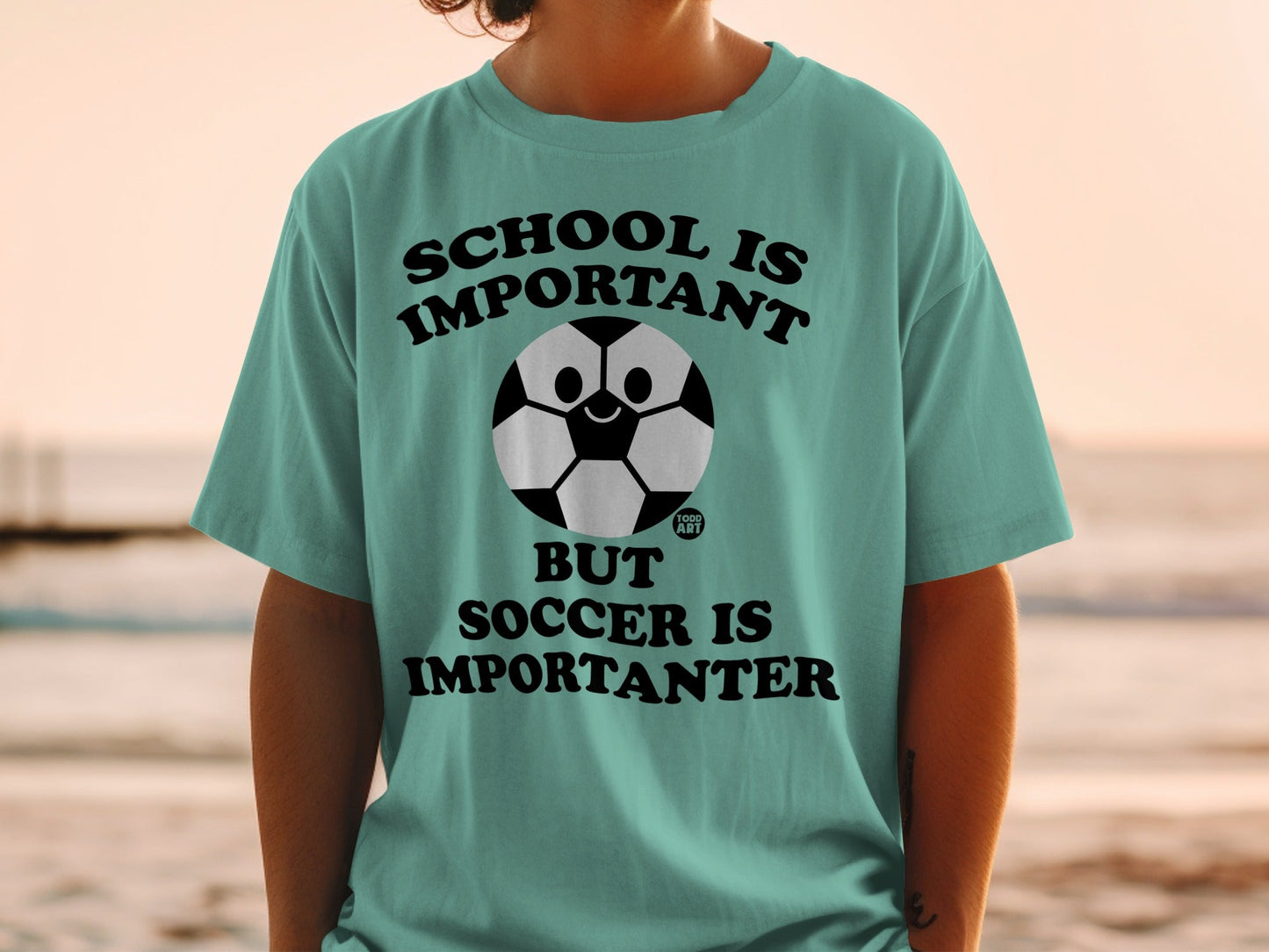 Teal t-shirt with cartoon soccer ball and text: School is important, but soccer is importanter.