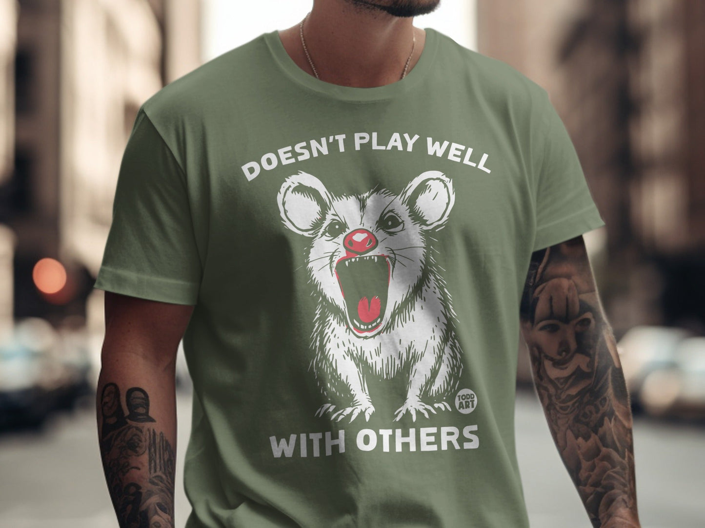 Olive green t-shirt with a cartoon opossum yelling "Doesn't Play Well With Others".