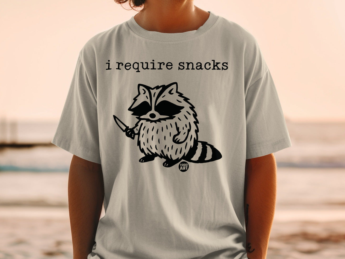 T-shirt featuring a raccoon holding a knife with text "I require snacks" and "1000 ART".