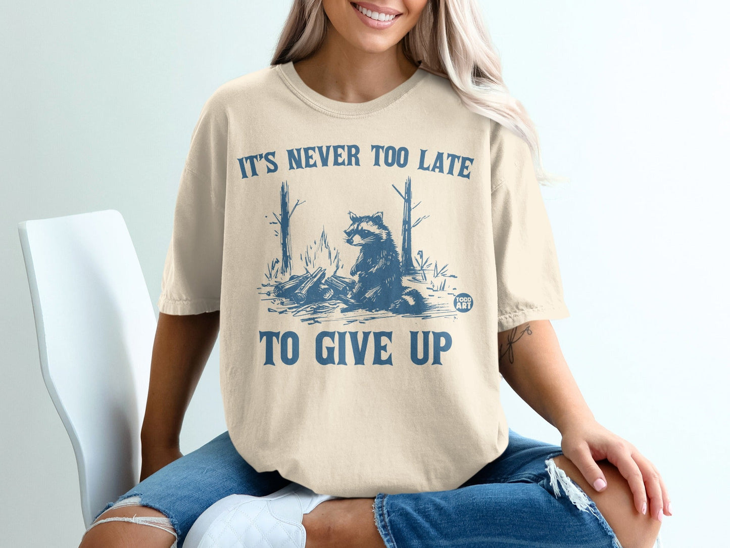 Cream t-shirt with raccoon graphic and "IT'S NEVER TOO LATE TO GIVE UP" text.