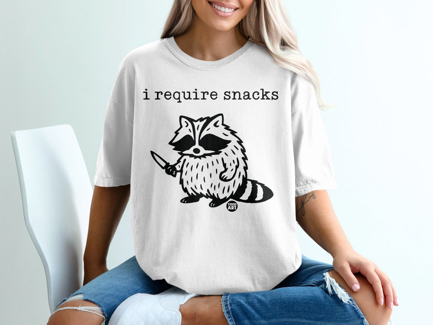 White t-shirt with a raccoon holding a knife and the text "I require snacks."