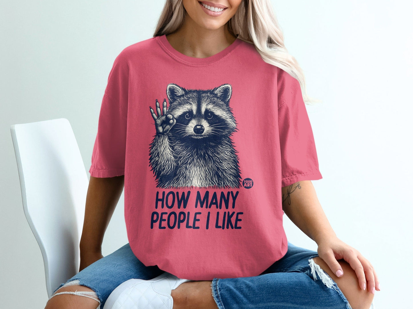 Pink t-shirt with raccoon graphic and text "HOW MANY PEOPLE I LIKE".