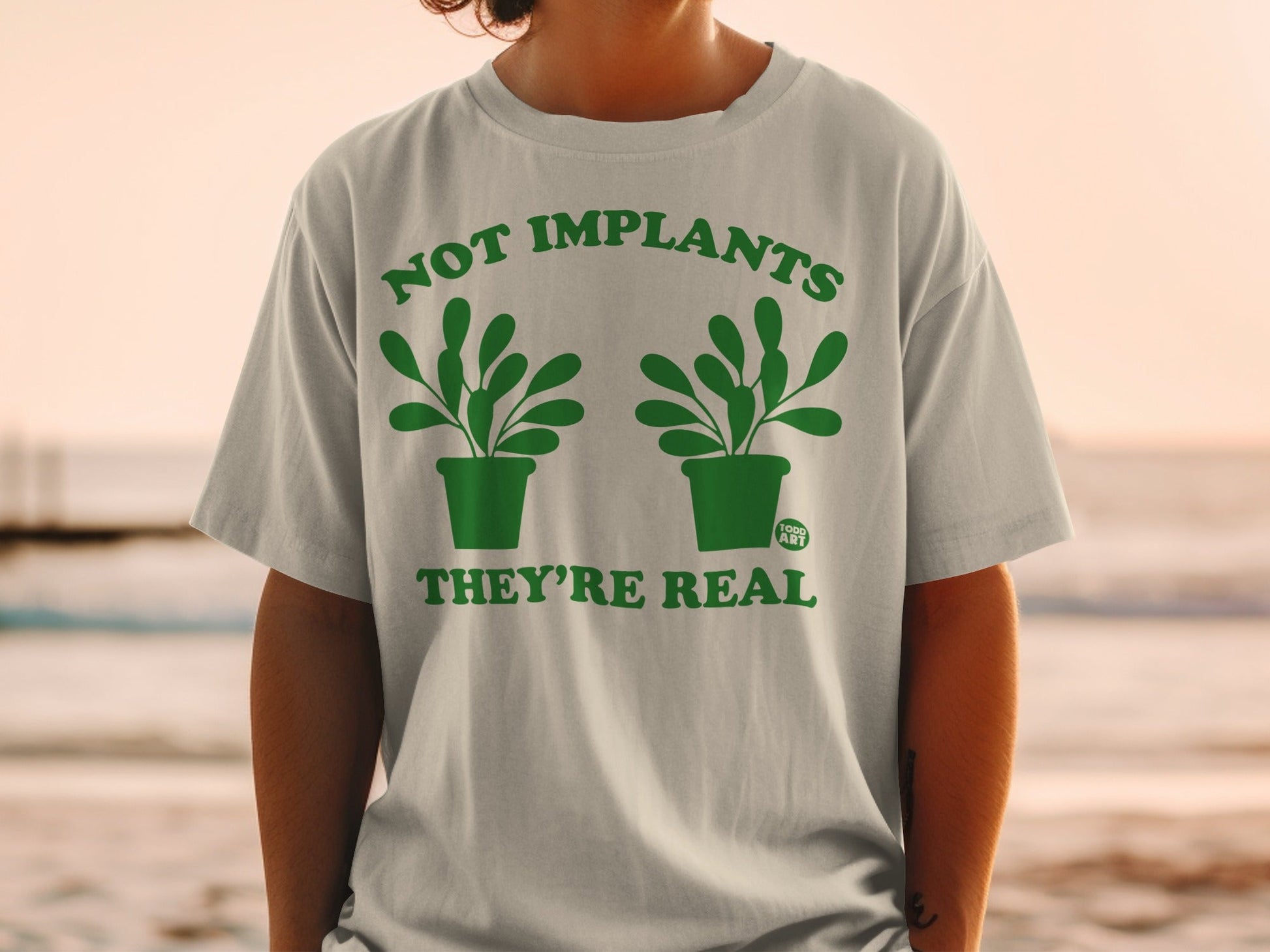 "Not implants, they're real" text on a grey t-shirt with green potted plants.