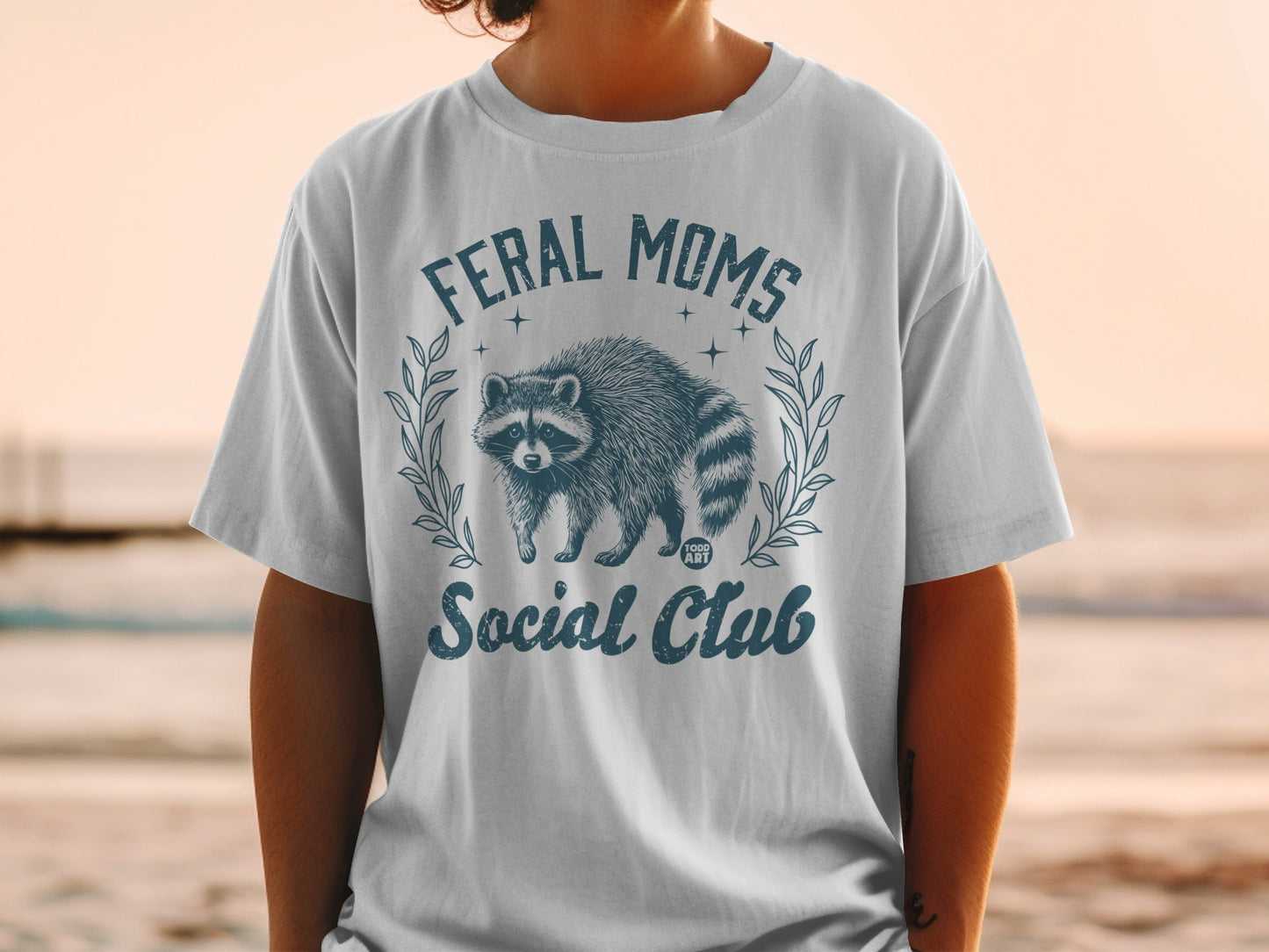 "Feral Moms Social Club" graphic on a white t-shirt featuring a raccoon.