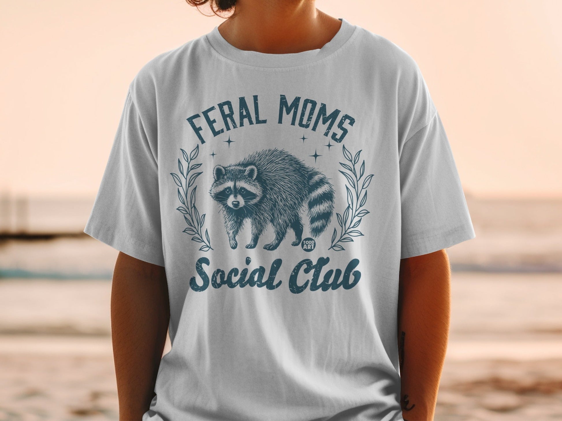 "Feral Moms Social Club" graphic on a white t-shirt featuring a raccoon.