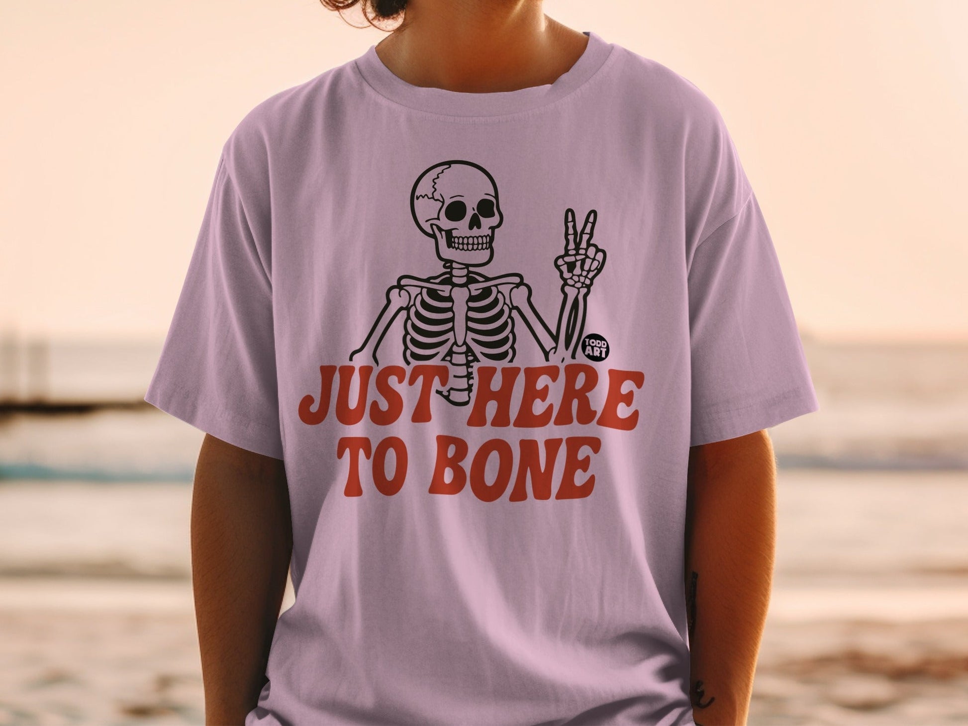 Skeleton graphic with text "JUST HERE TO BONE" on a light purple t-shirt.