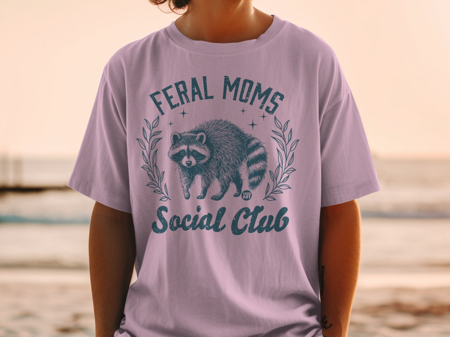 Feral Moms Social Club text with raccoon illustration on a purple t-shirt.