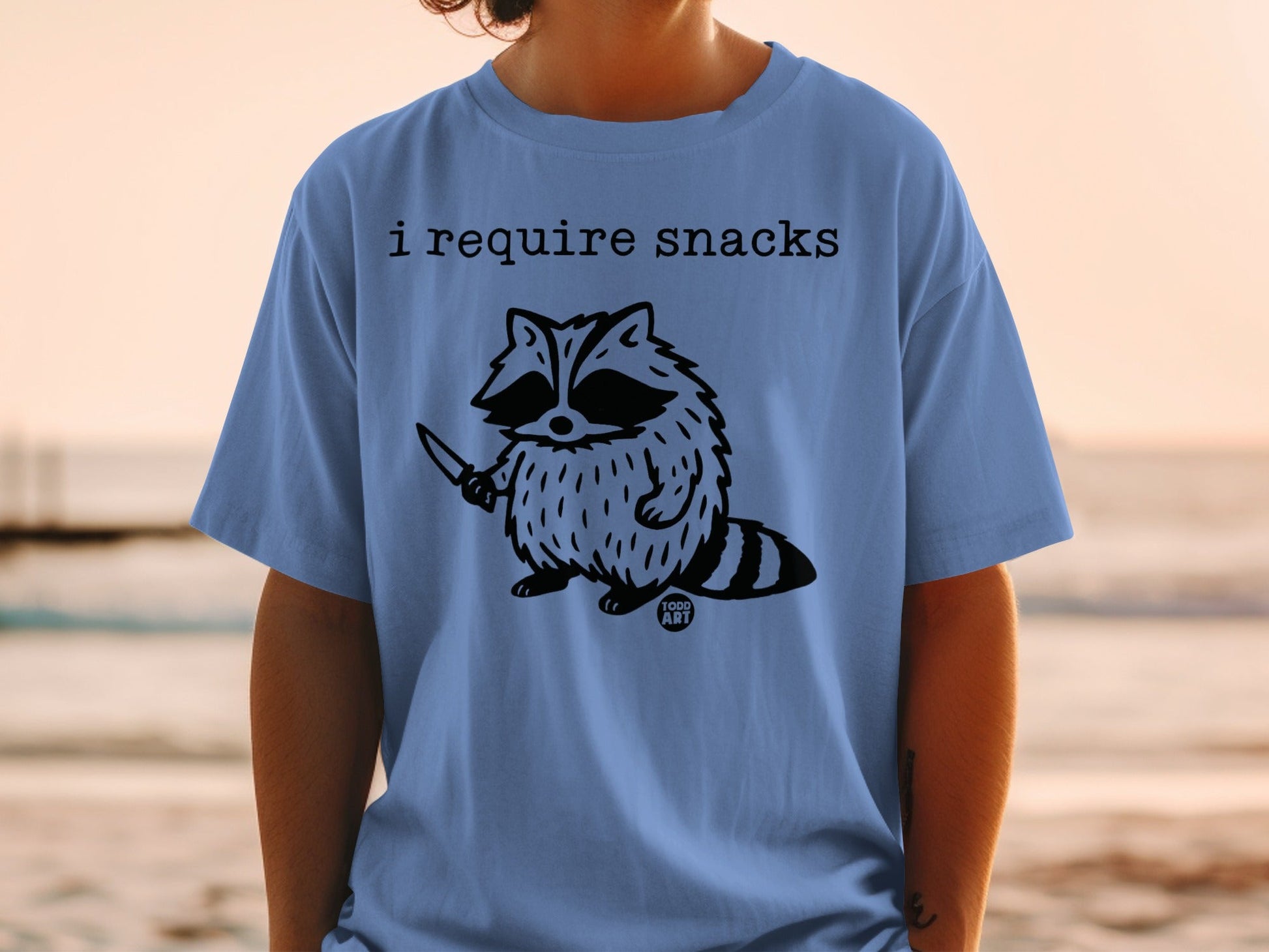Blue t-shirt with "I require snacks" and a raccoon holding a knife.