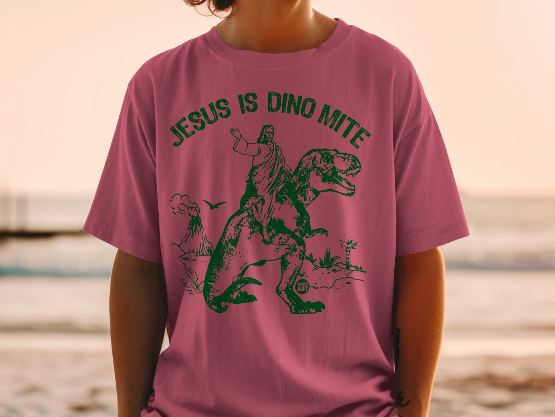 Pink t-shirt with green graphic of Jesus riding a dinosaur.