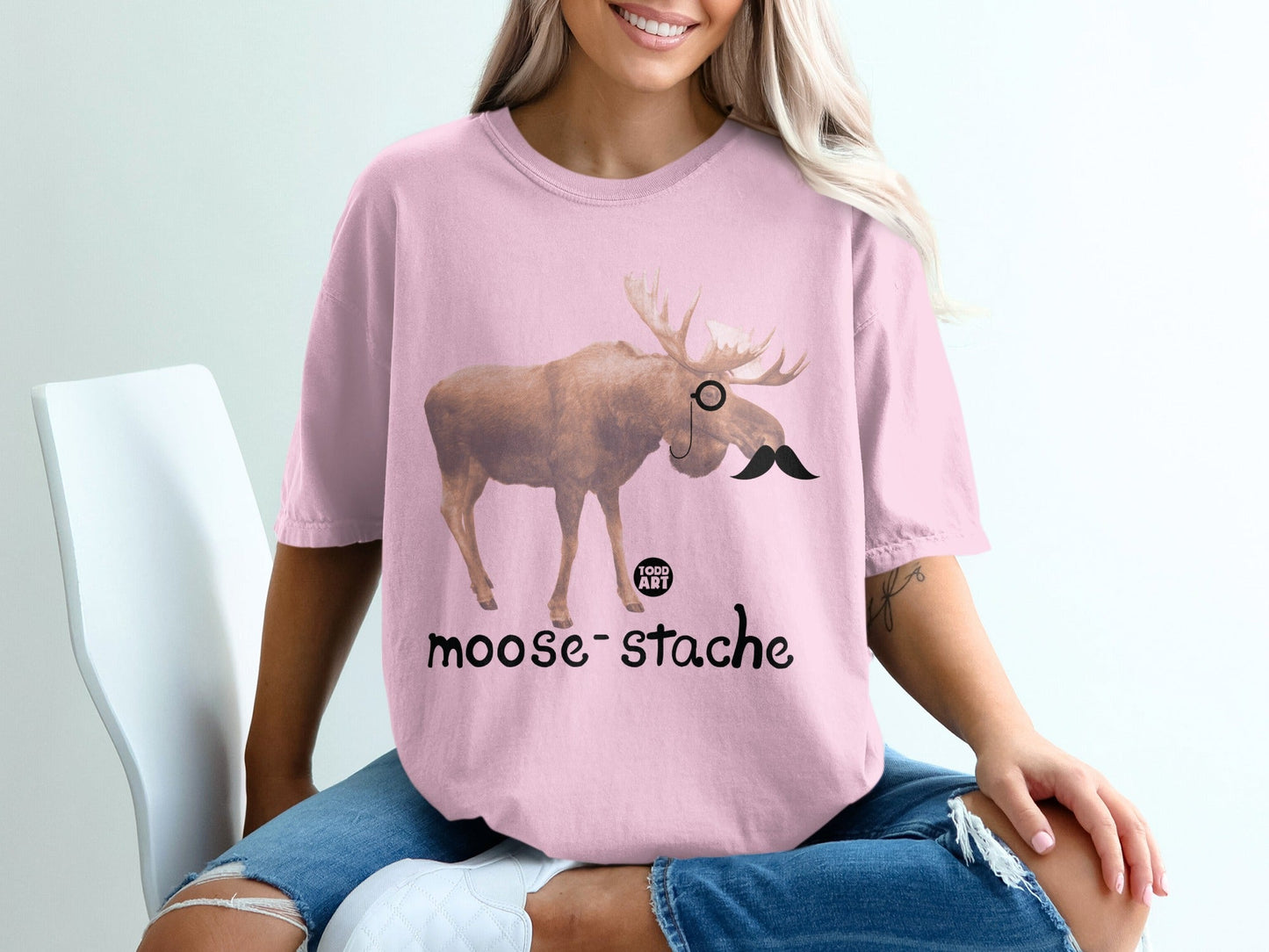 Pink t-shirt with a moose wearing a monocle and mustache, text reads "moose-stache".