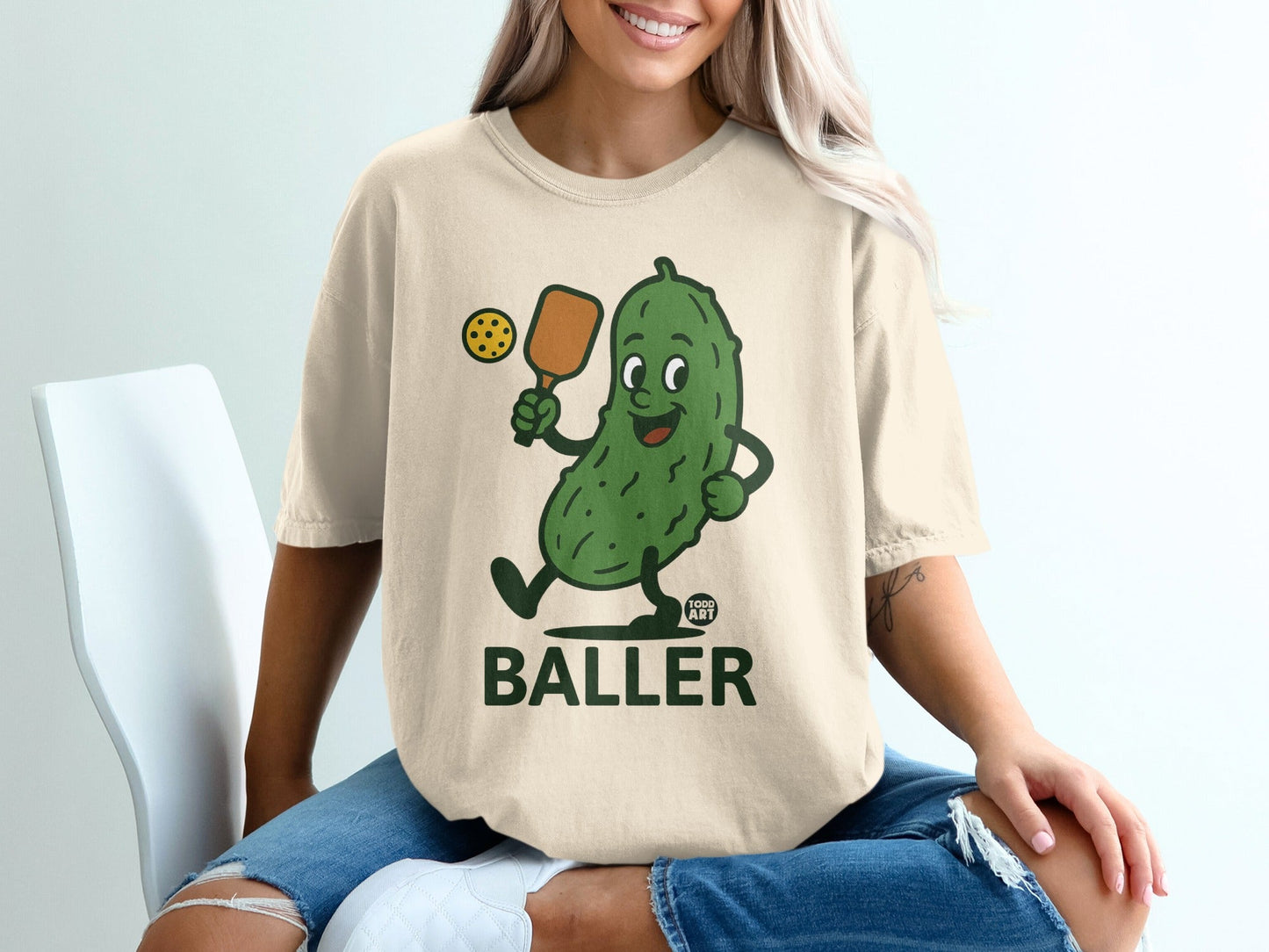Cartoon pickle playing pickleball, "BALLER" text on beige t-shirt.