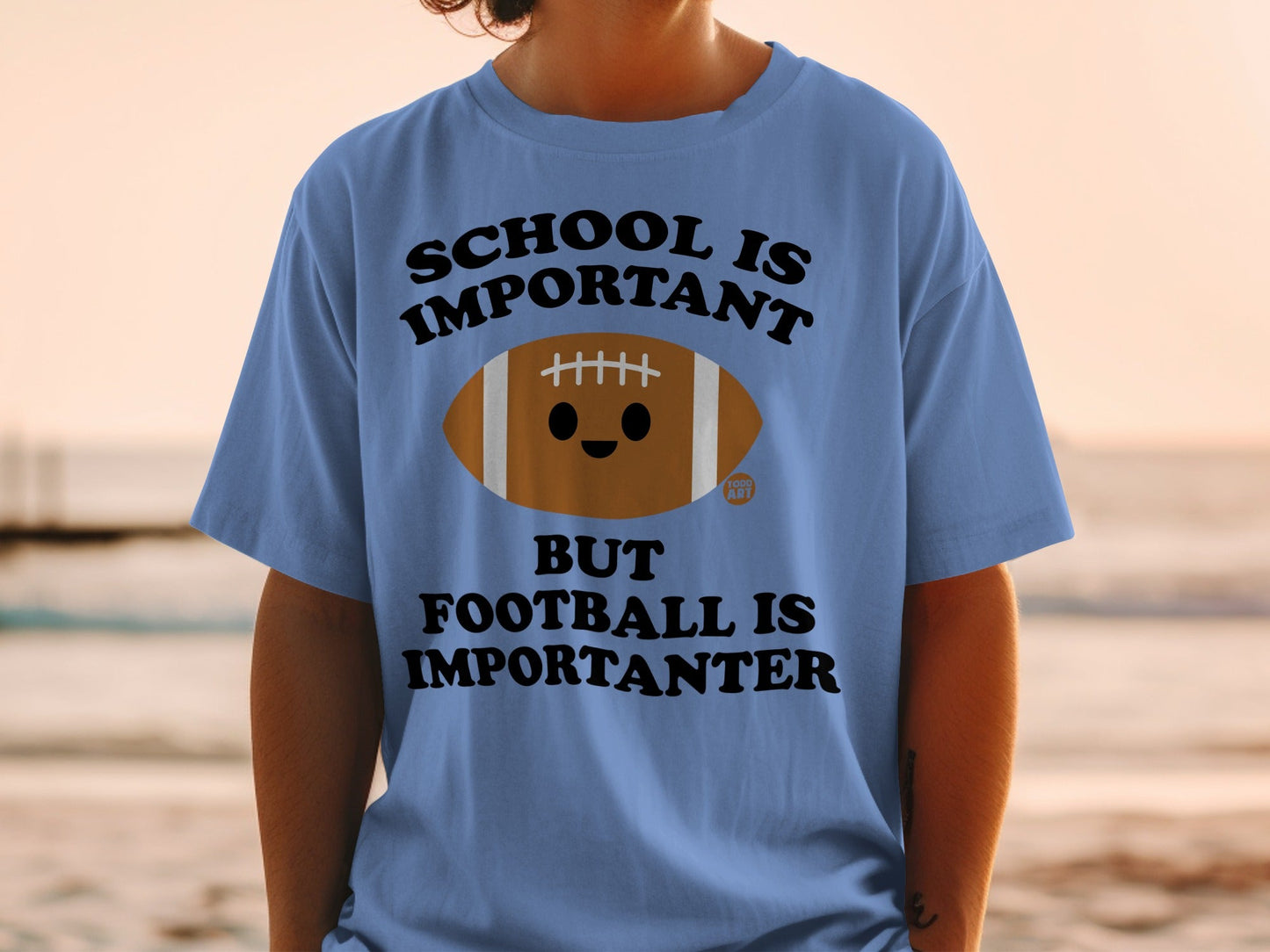 Light blue t-shirt with cartoon football and text "School is Important but Football is Importanter".
