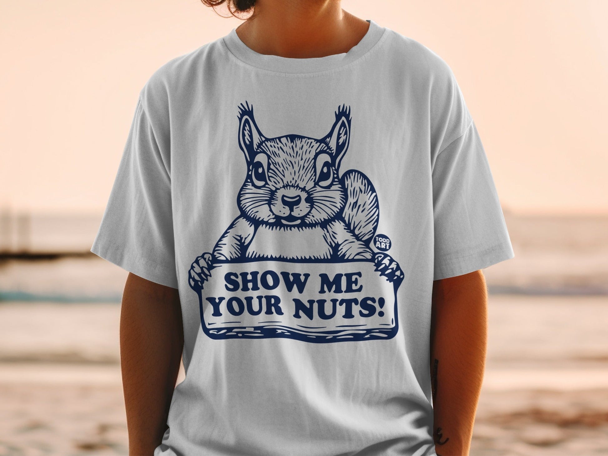 White t-shirt with a cartoon squirrel holding a sign that says "SHOW ME YOUR NUTS!"