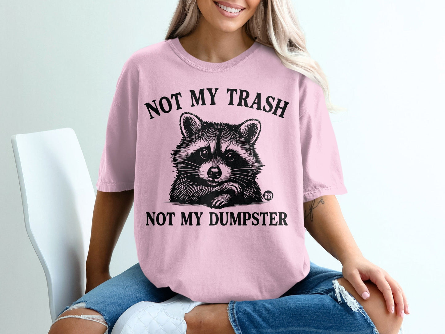 Pink t-shirt with raccoon graphic and text "NOT MY TRASH NOT MY DUMPSTER".