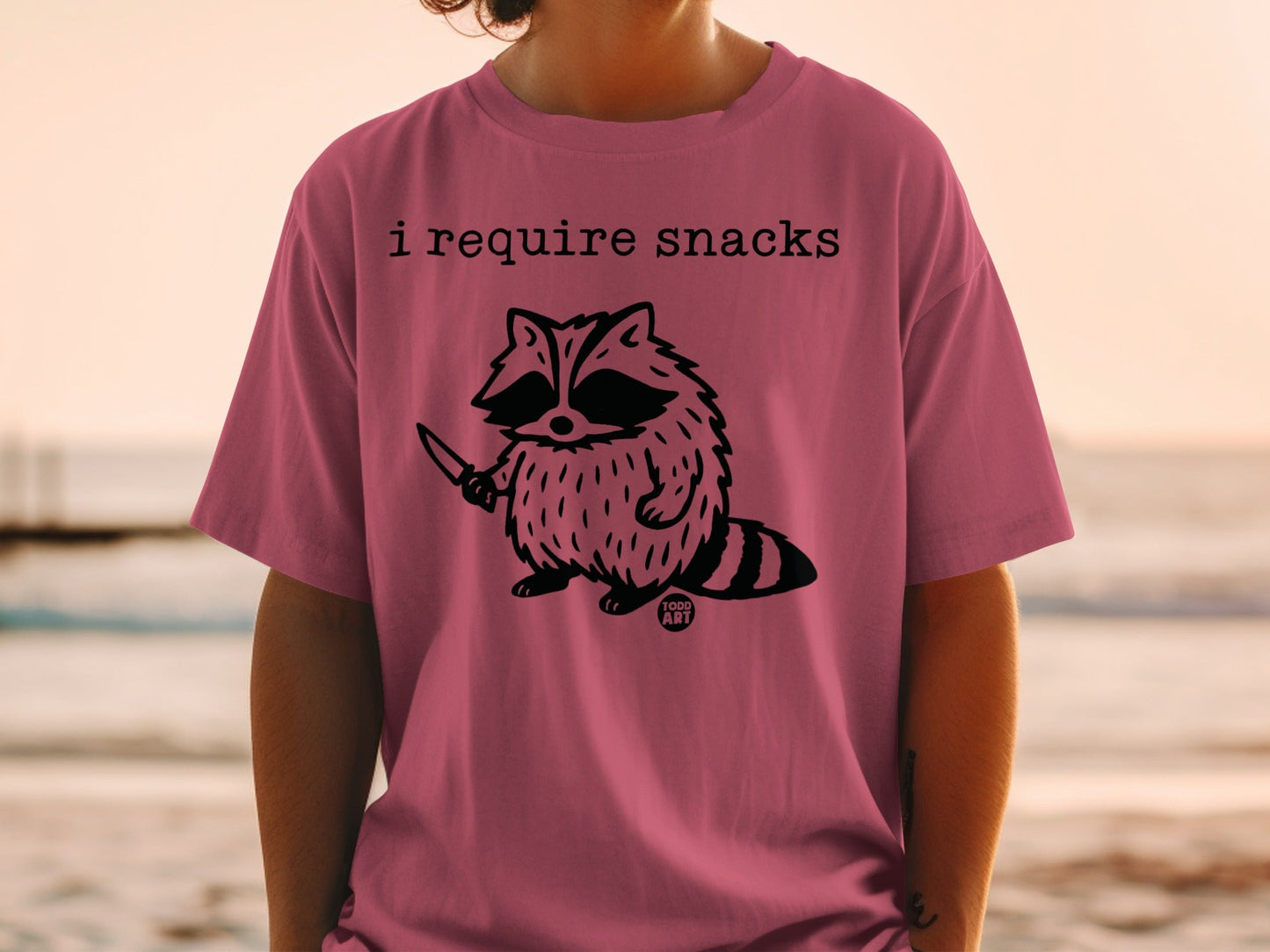 T-shirt with a raccoon holding a knife and the text "I require snacks".
