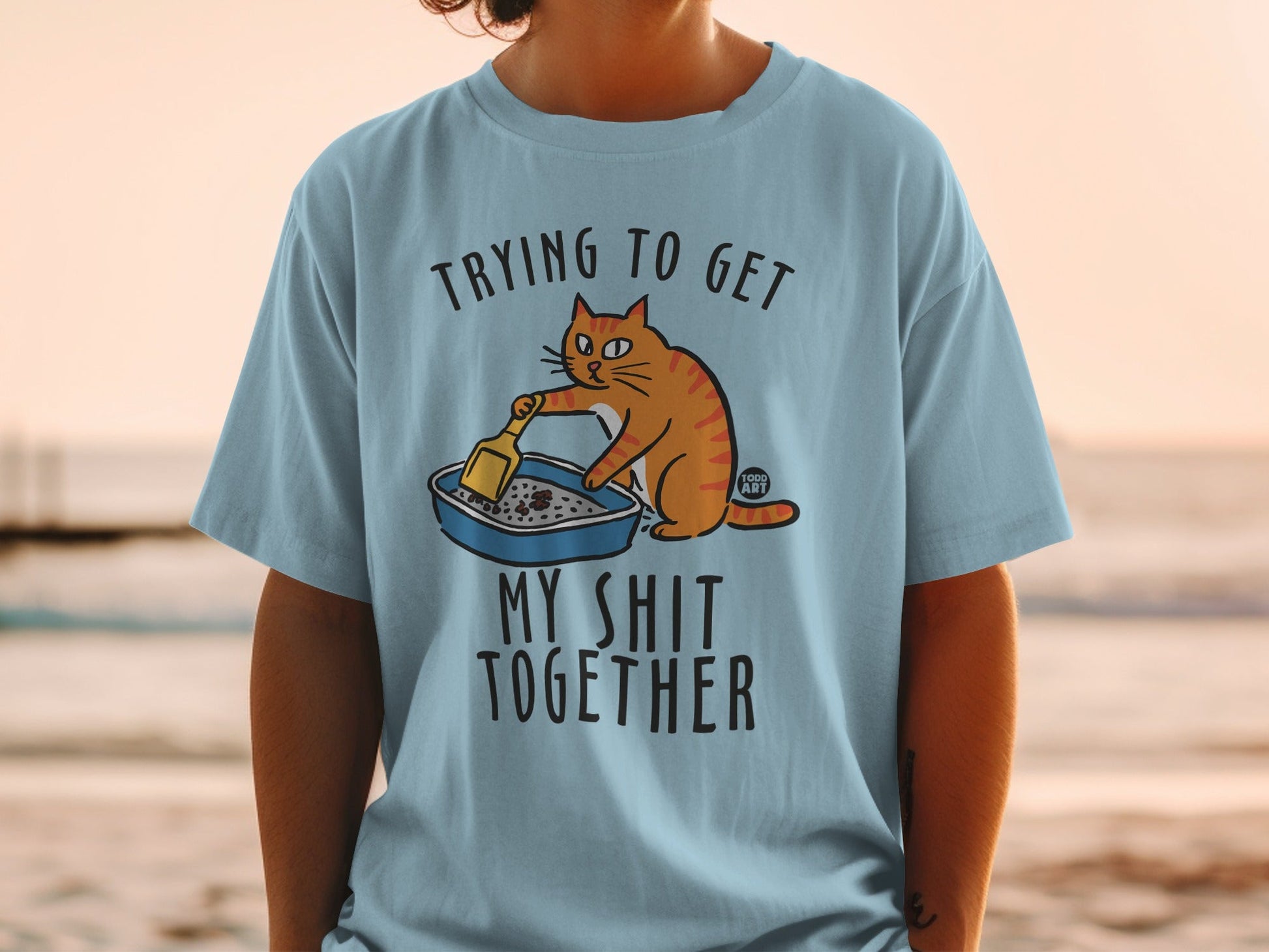 Light blue t-shirt with orange cat cleaning litter box, text "Trying to get my shit together".