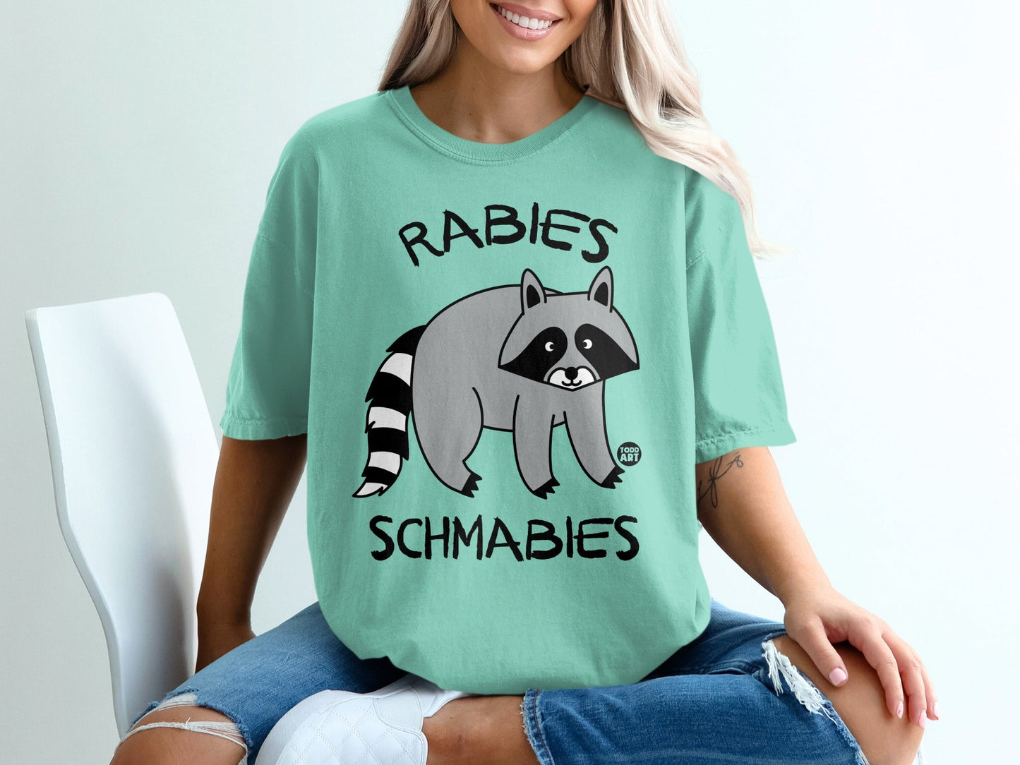 Raccoon graphic with text "RABIES SCHMABIES" on a mint green t-shirt.