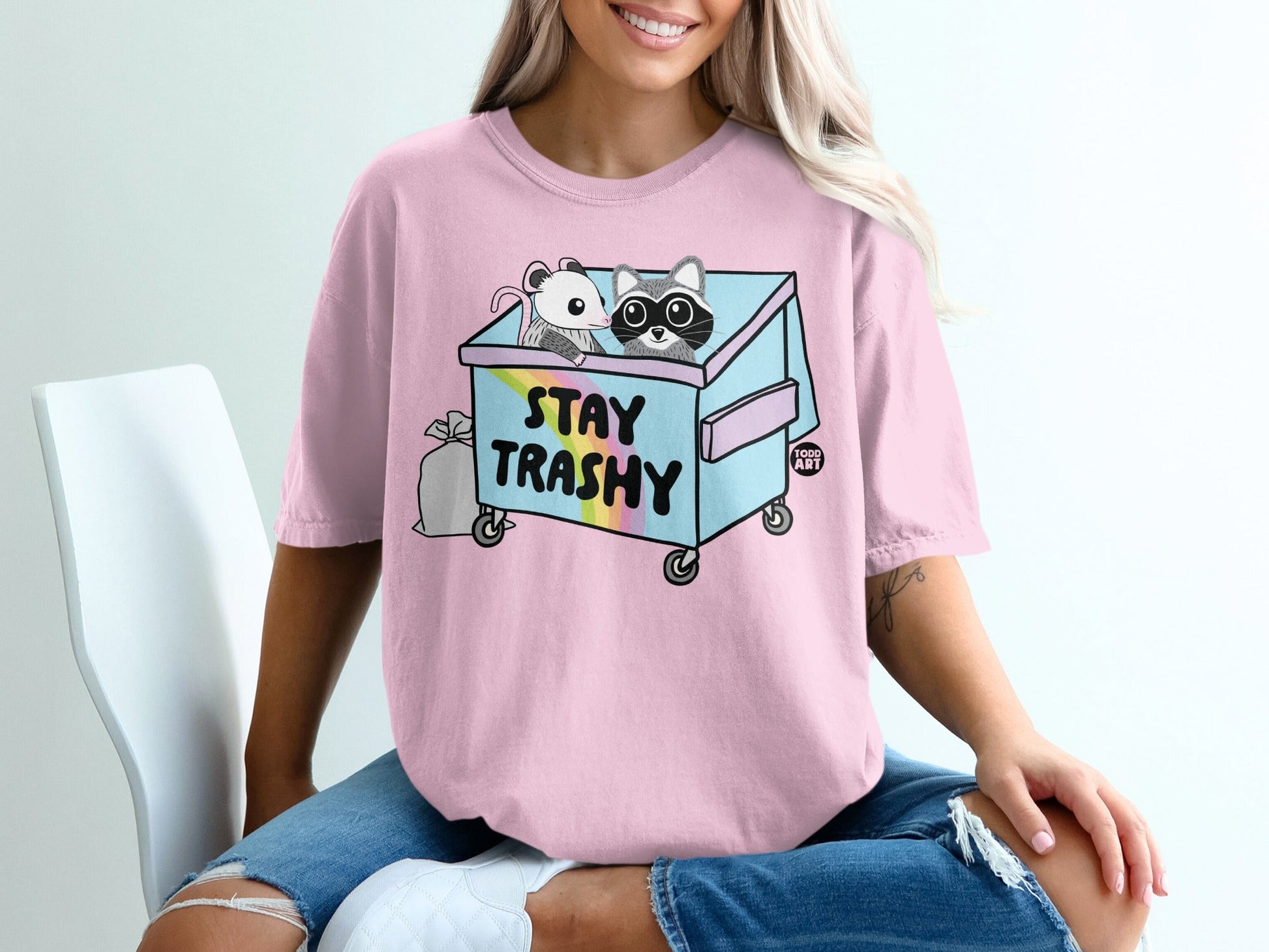 Pink t-shirt with a cartoon raccoon and mouse in a blue dumpster with rainbow accents.
