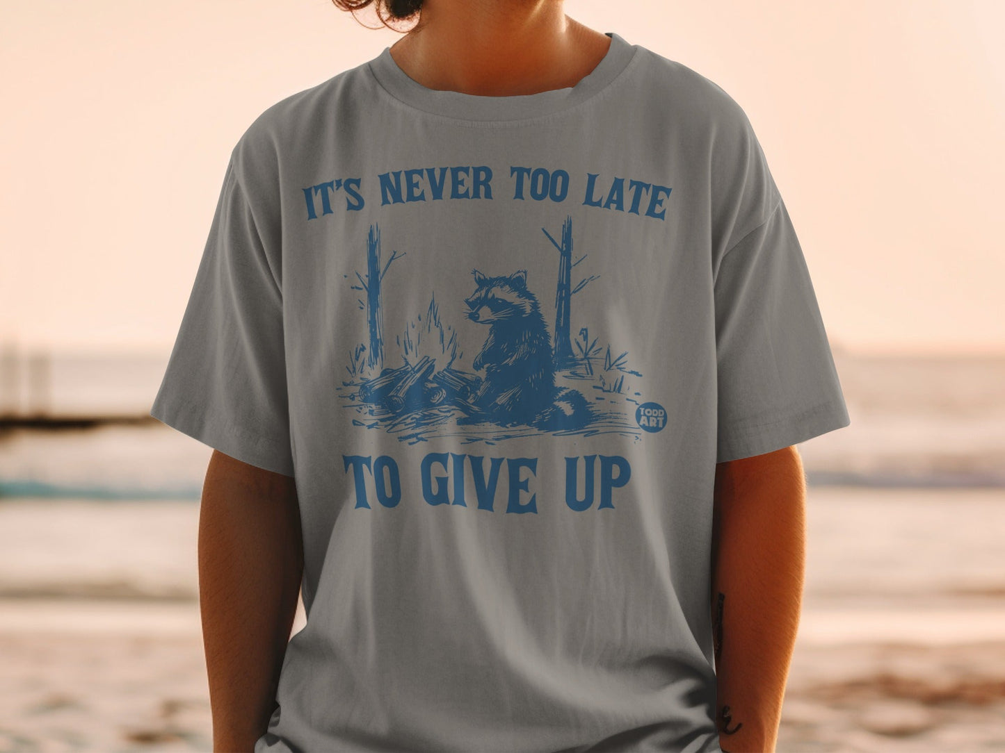 T-shirt with blue graphic of a raccoon by a campfire with text "IT'S NEVER TOO LATE TO GIVE UP".