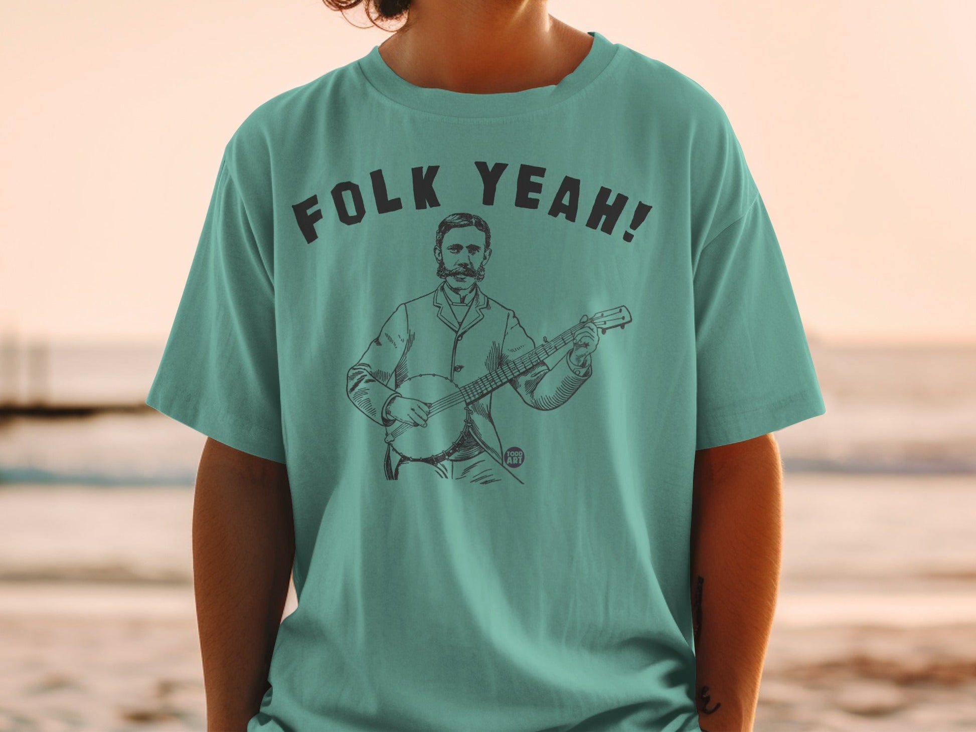 Teal t-shirt with "Folk Yeah!" text above a line drawing of a man playing a banjo.