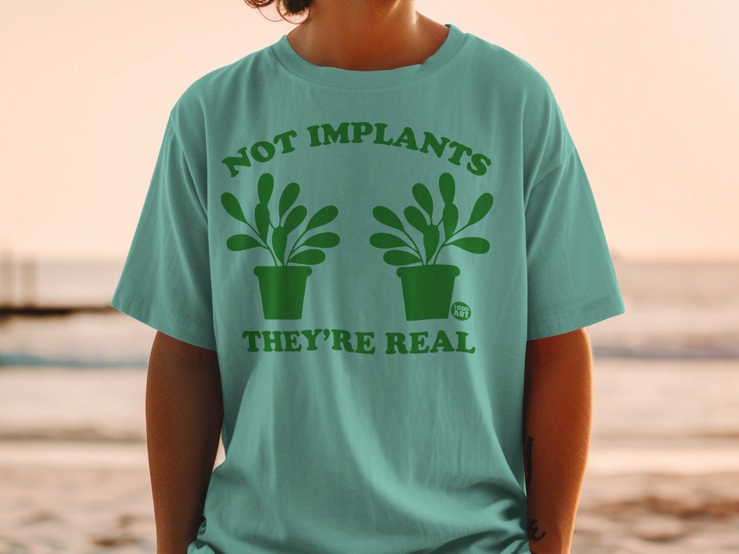 Teal t-shirt with green text and plants: "NOT IMPLANTS THEY'RE REAL".