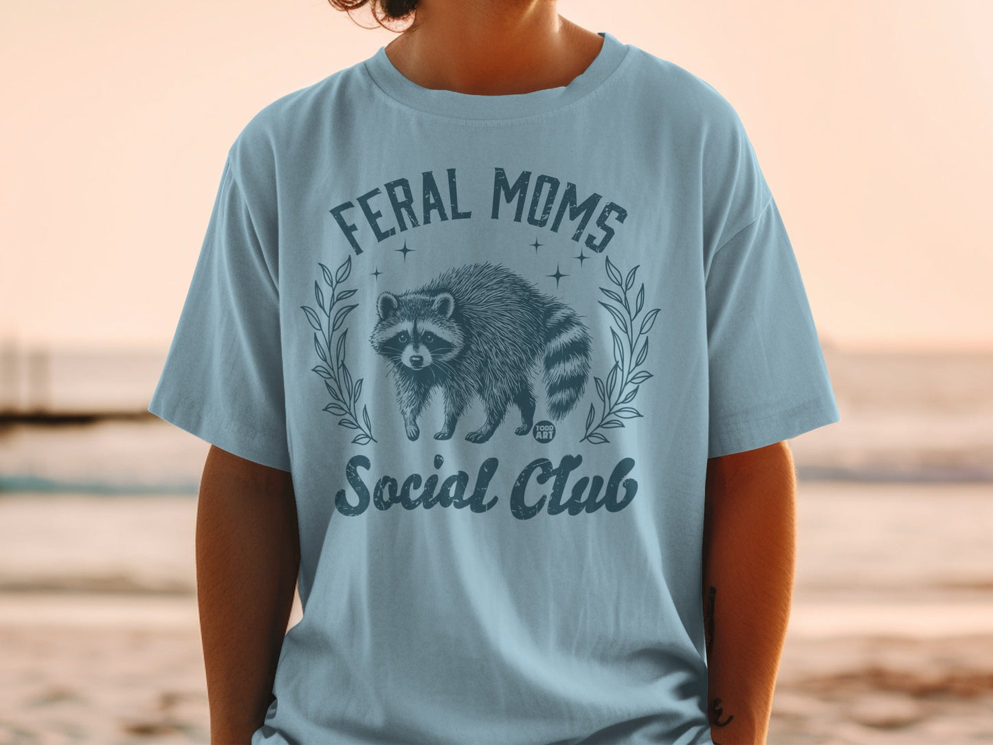 Light blue t-shirt with "Feral Moms Social Club" and raccoon graphic.